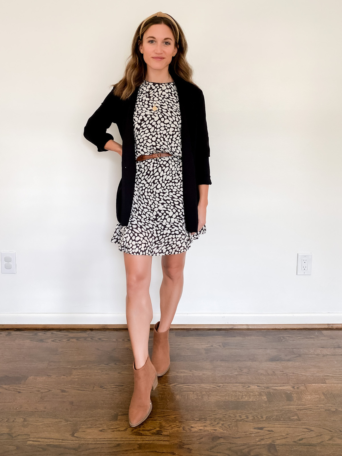 ashley wearing a black and white patterned dressed belted with a skinny brown belt, a black blazer, and cognac ankle boots