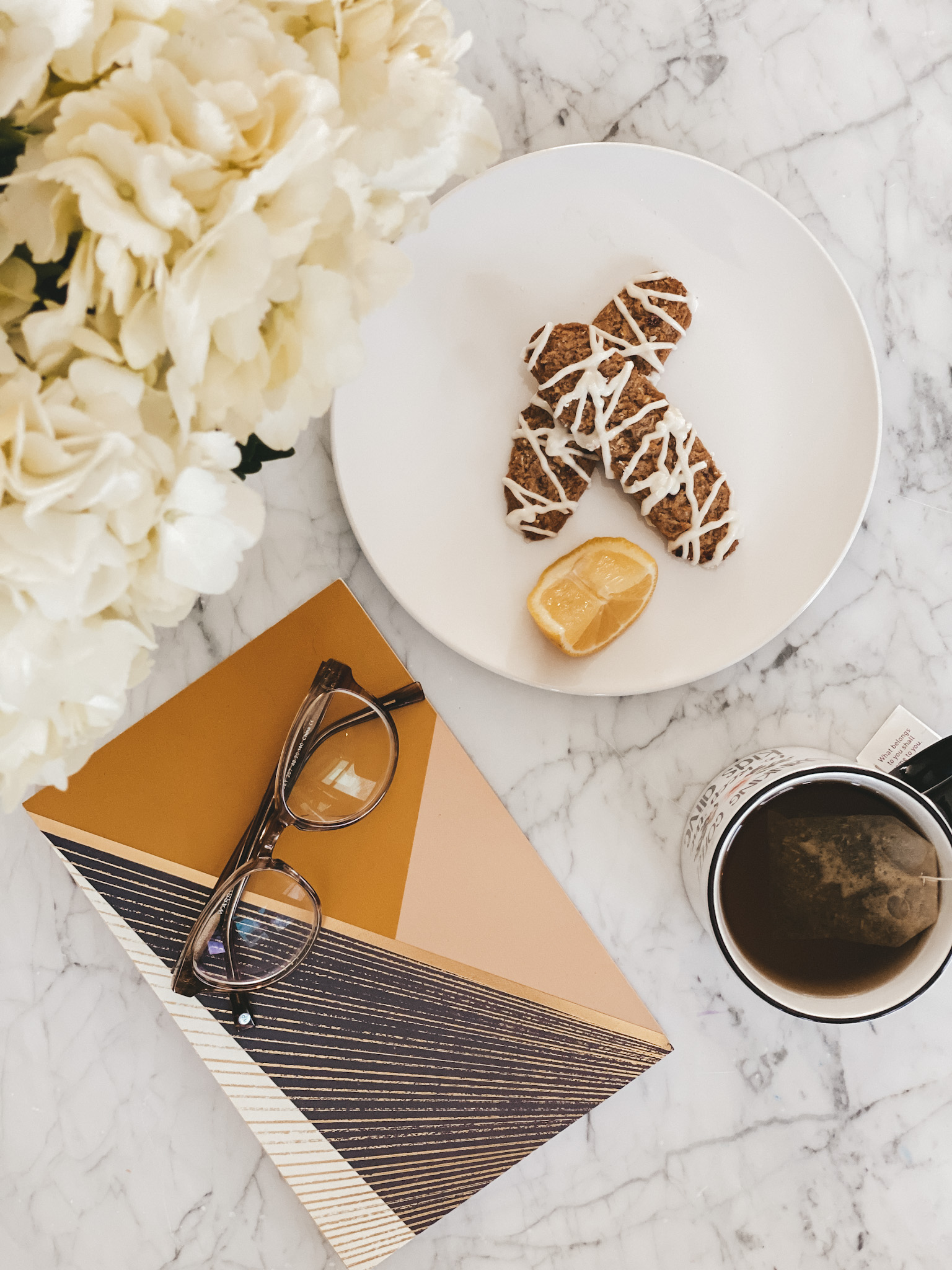 white hydrangeas next to a notebook with glasses set on top, a white plate with 2 iced cookies and a slice of lemon, a mug of tea on a marble table