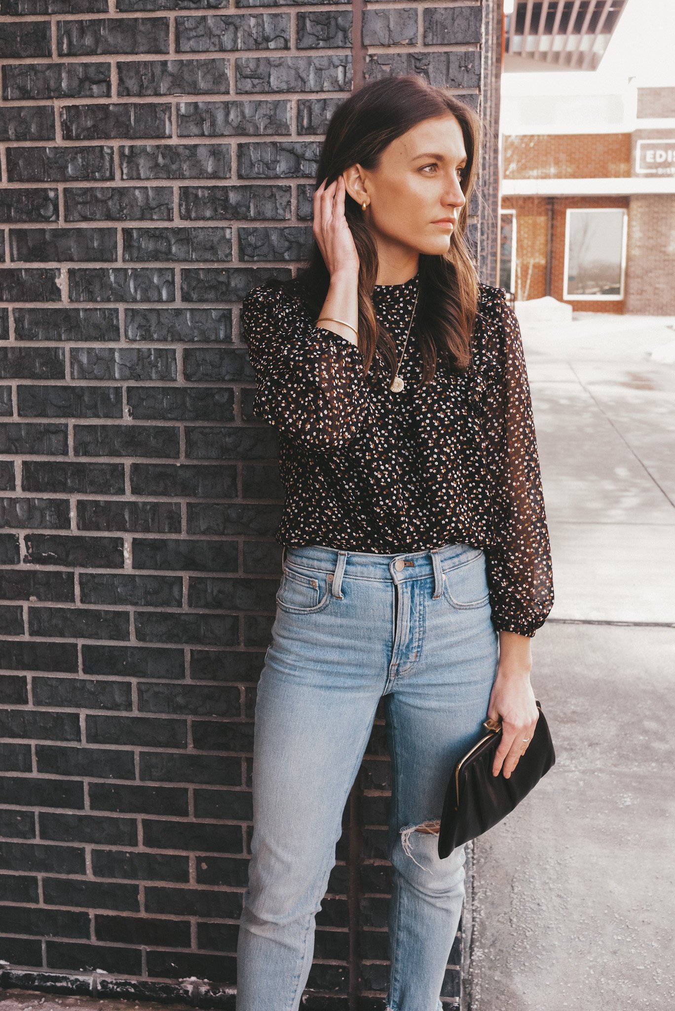 ashley wearing simple jewelry including gold hoop earrings, a pendant necklace and gold bangle with a black floral top and light wash distressed jeans from Madewell standing in front of a gray brick wall