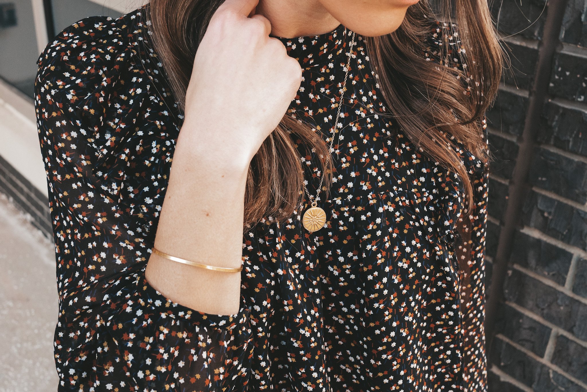 ashley wearing simple jewelry including gold hoop earrings, a pendant necklace and gold bangle with a black floral top and light wash distressed jeans from Madewell standing in front of a gray brick wall