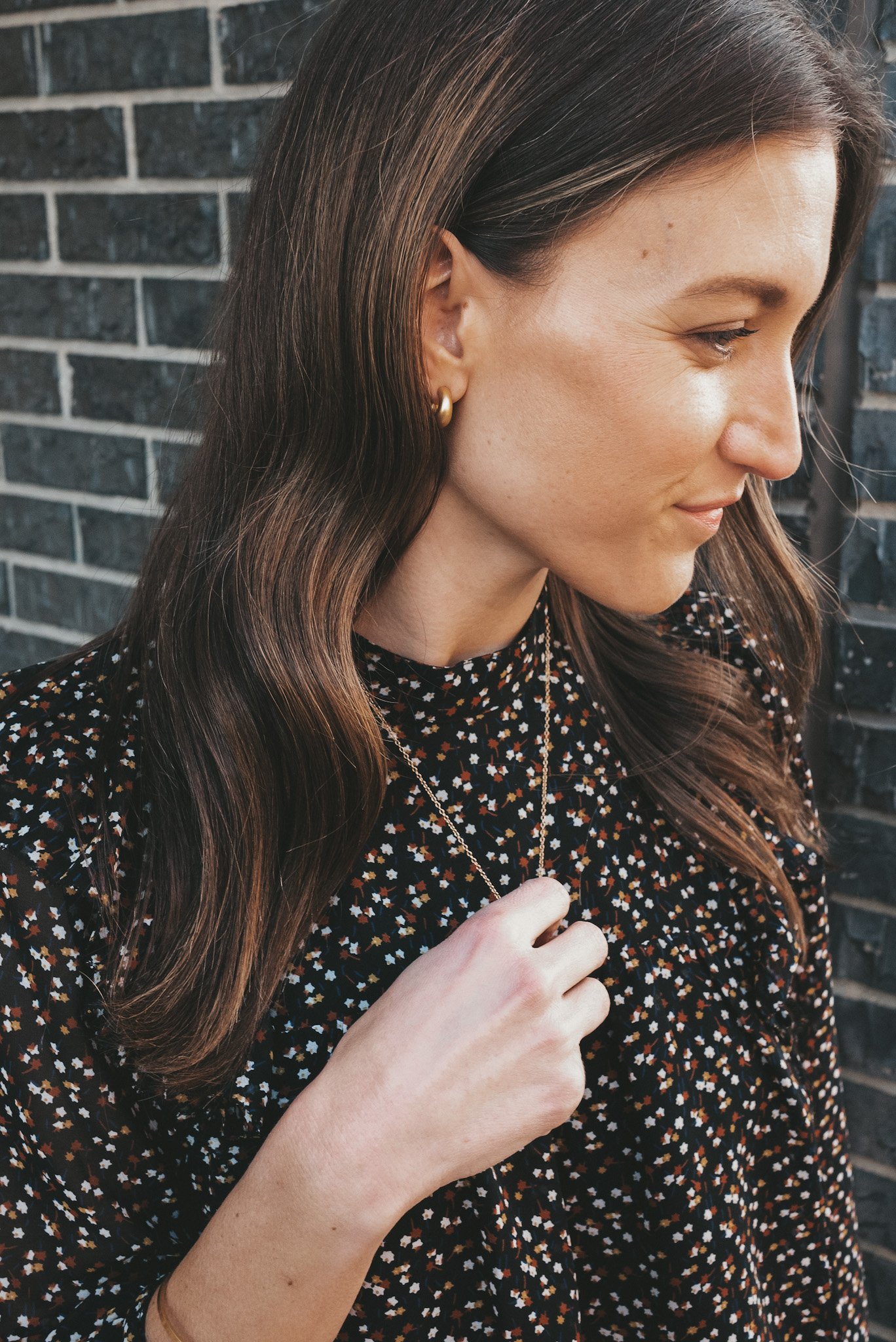 ashley wearing simple jewelry including gold hoop earrings, a pendant necklace and gold bangle with a black floral top