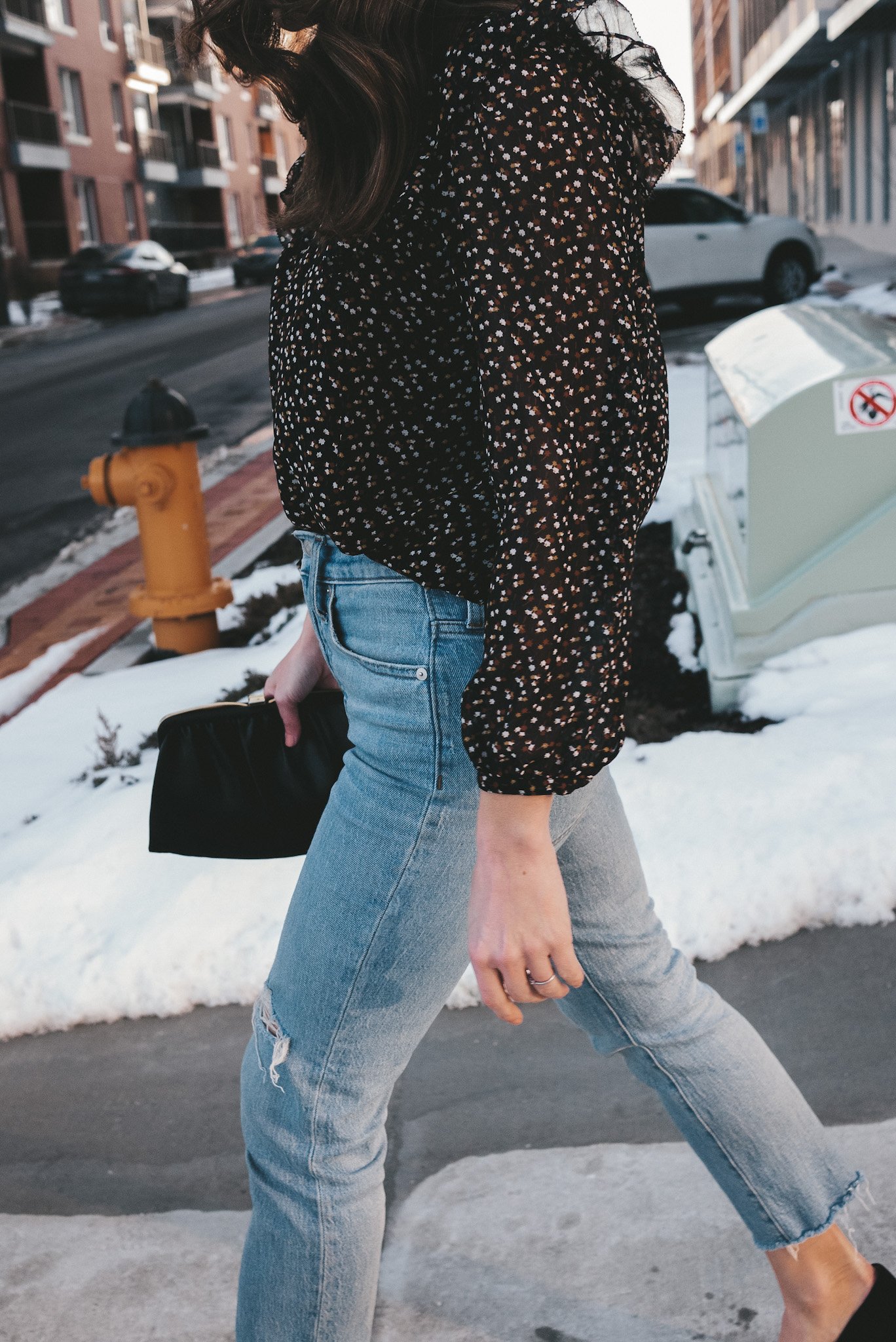 ashley wearing a black floral blouse and light wash distressed jeans from Madewell, carrying a black clutch and walking along a sidewalk