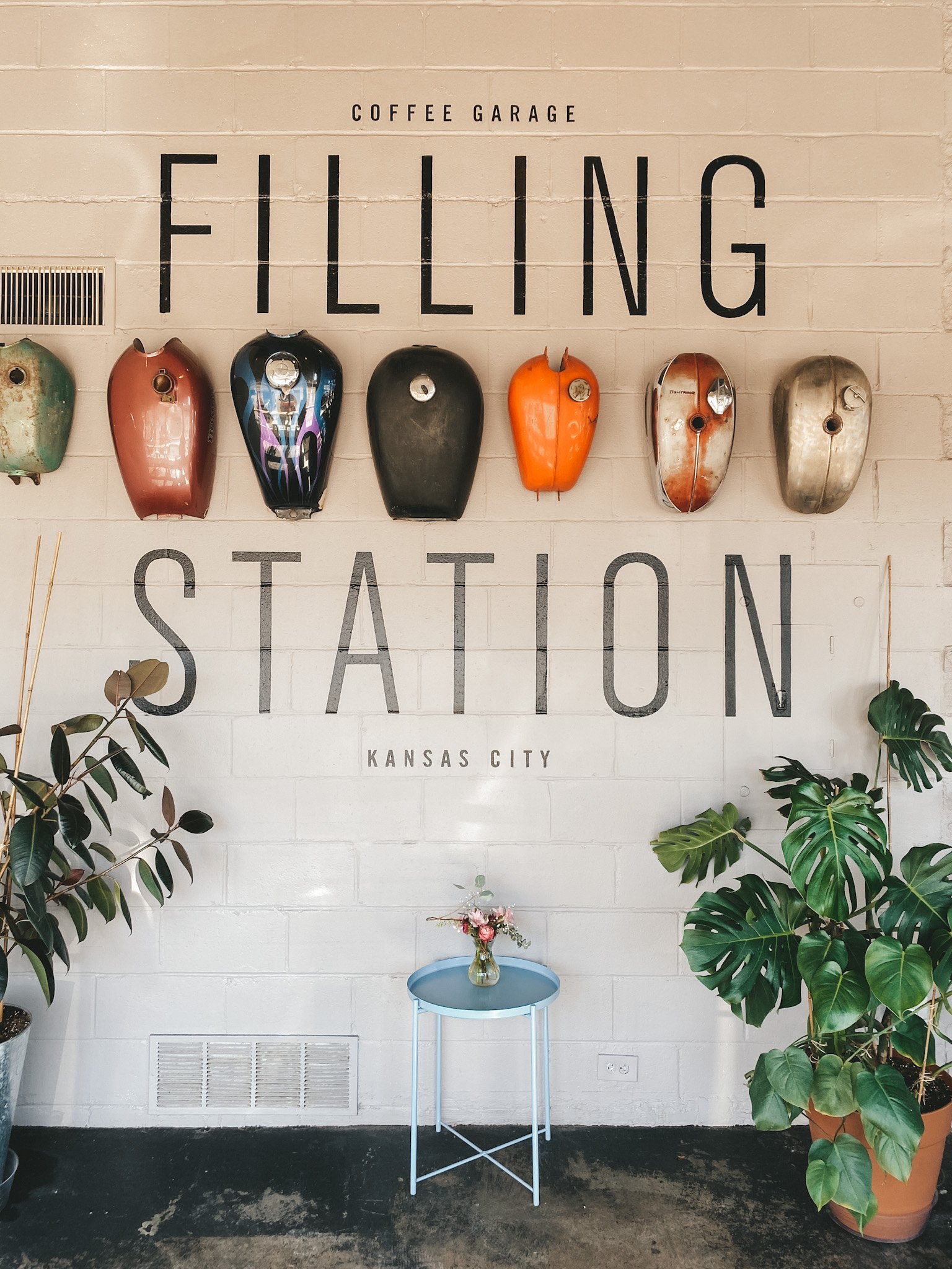 picture of The Filling Station Coffee Shop, bike helmets hanging on the wall, a small table, and 2 plants