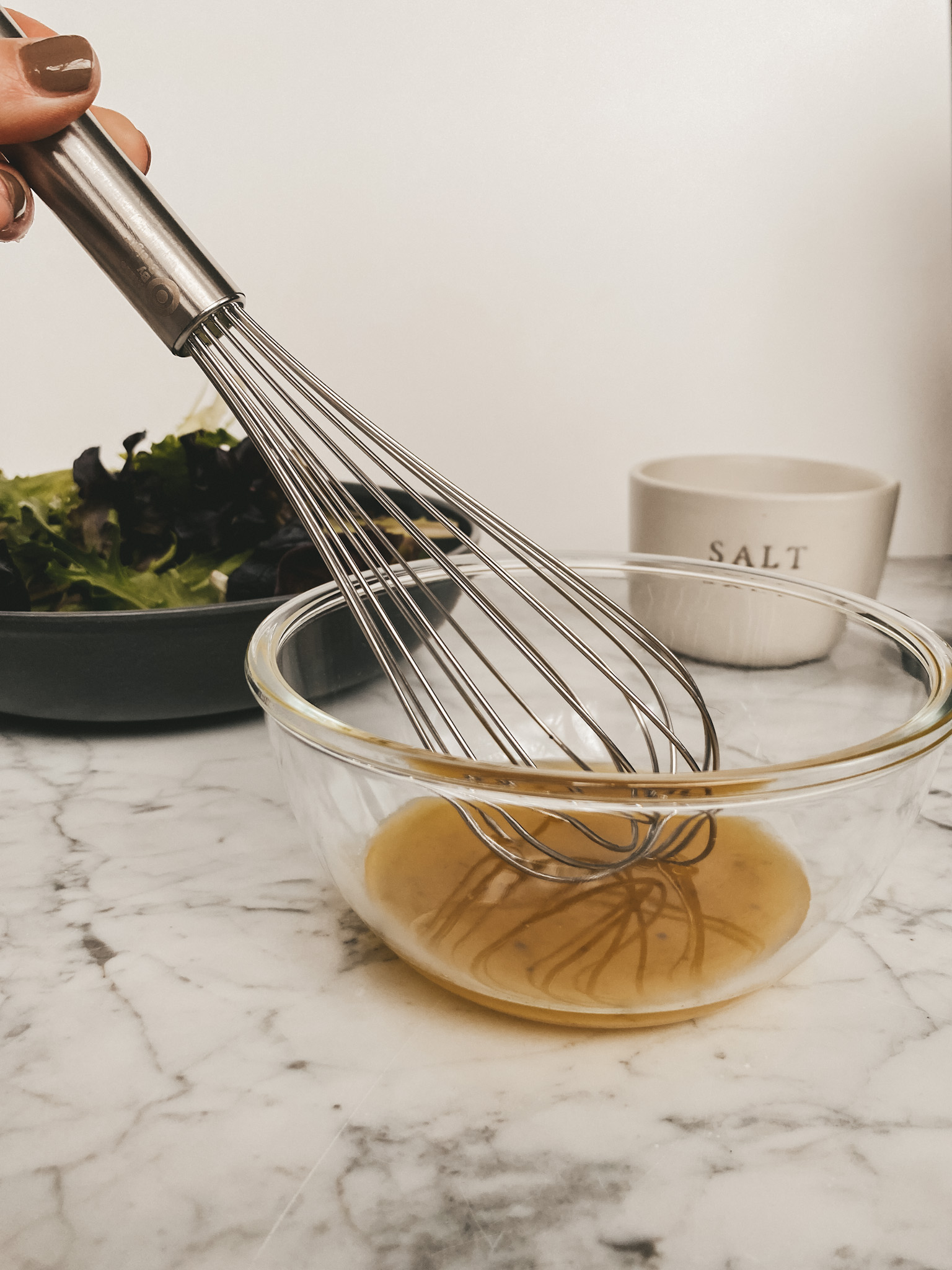 a bowl of lettuce, a bowl of salad dressing, a whisk and a ceramic container of salt
