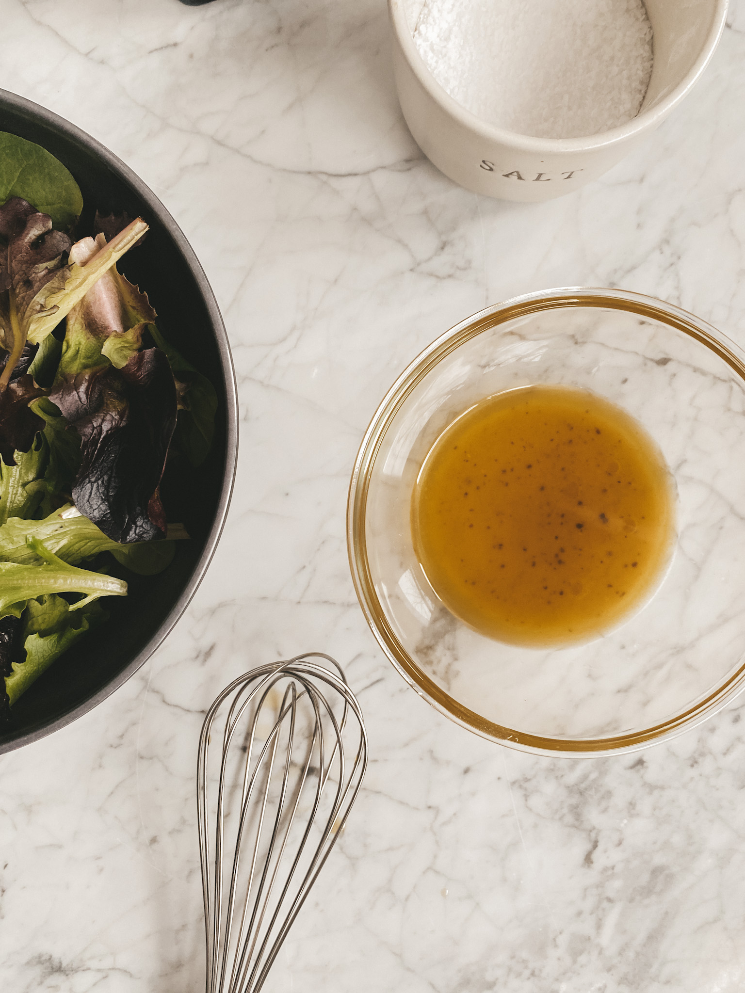 a bowl of lettuce, a bowl of salad dressing, a whisk and a ceramic container of salt