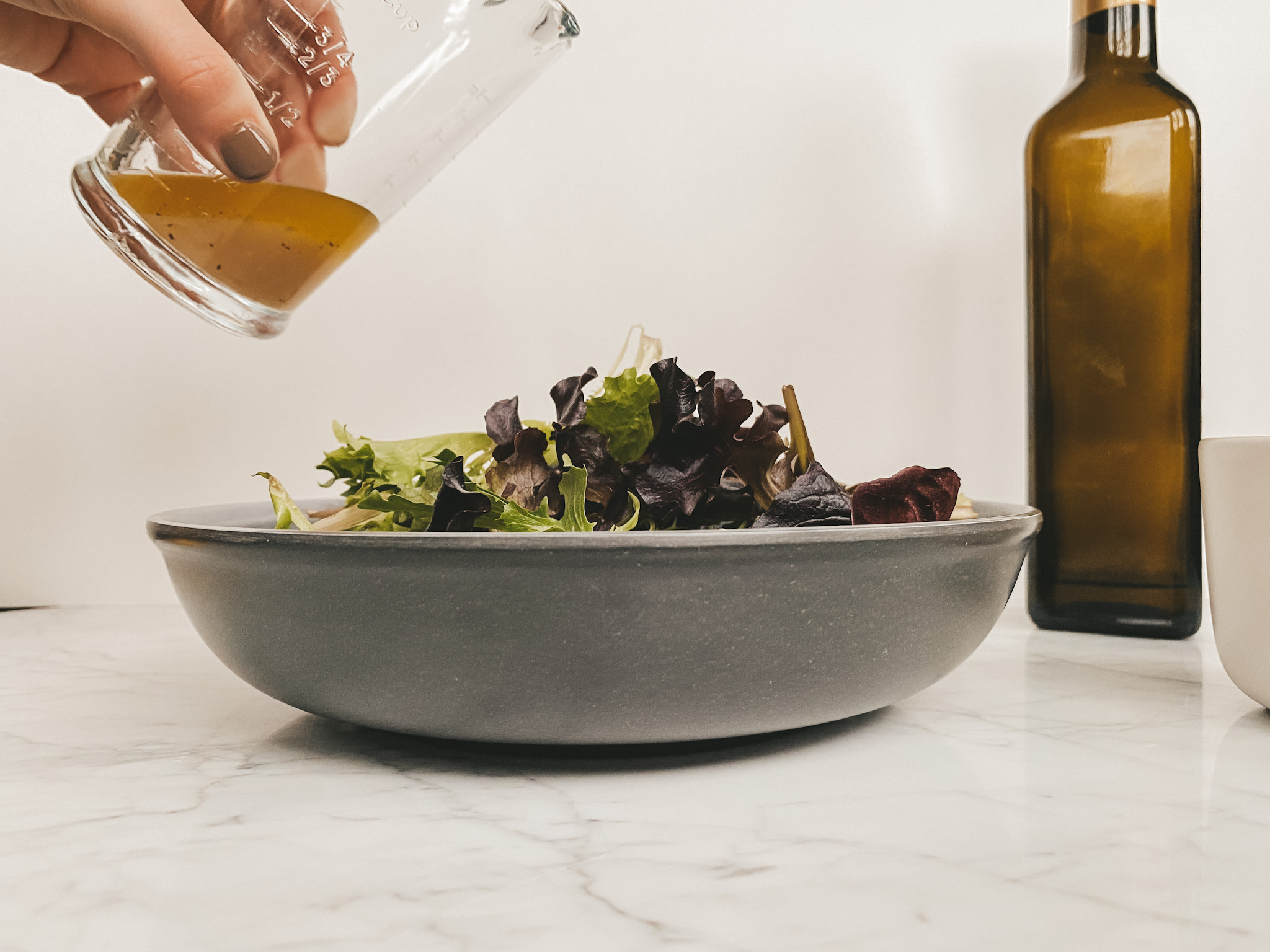 A gray bowl with mixed greens, a hand pouring dressing from a measuring cup over the salad