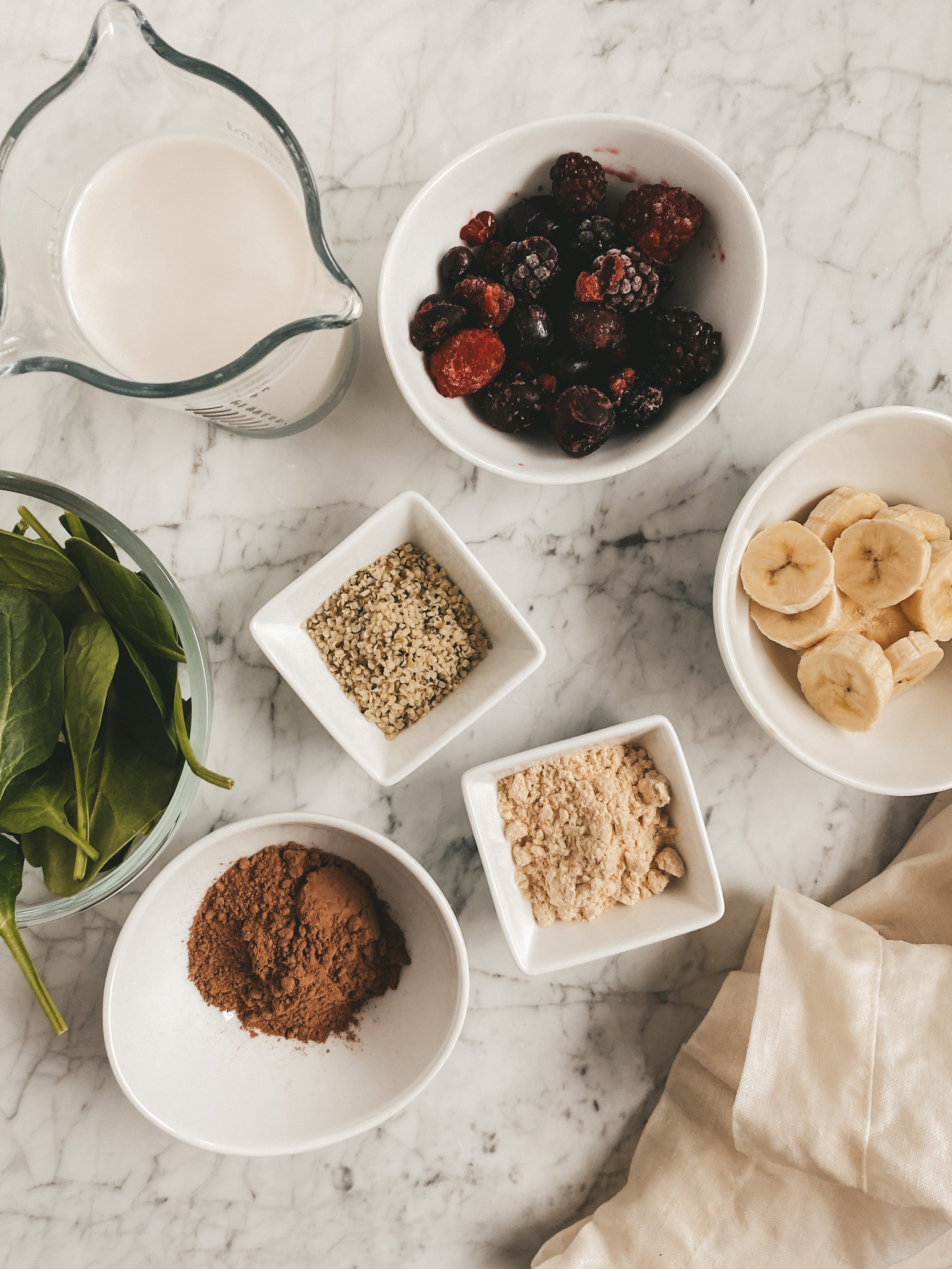assorted bowls on a marble table with smoothie ingredients: spinach, almond milk, banana, cherries, hemp seeds, protein powder, and cocoa powder
