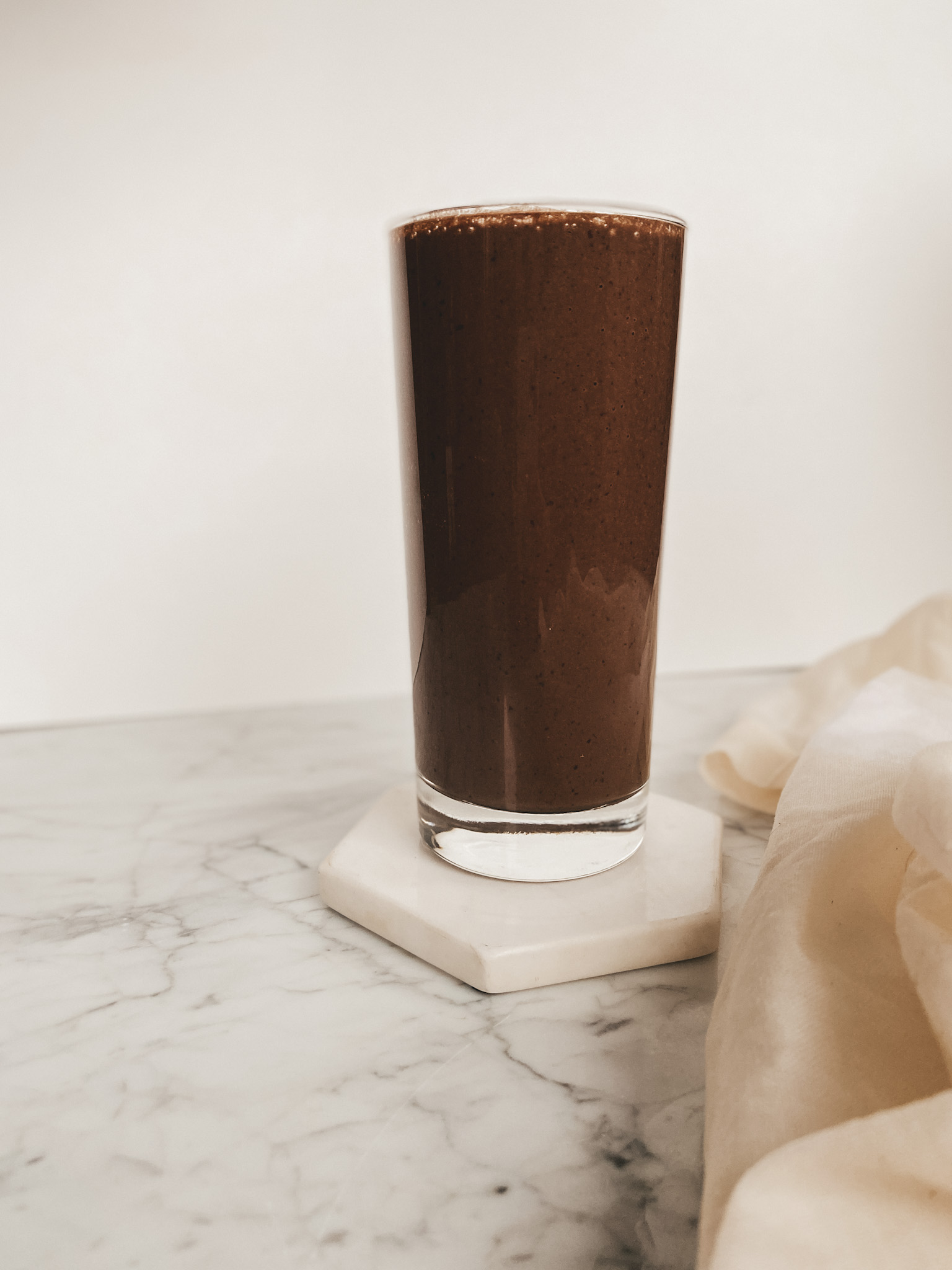 a purple smoothie in a clear glass on a marble coaster set on a marble table with a cream colored linen tea towel and a white background