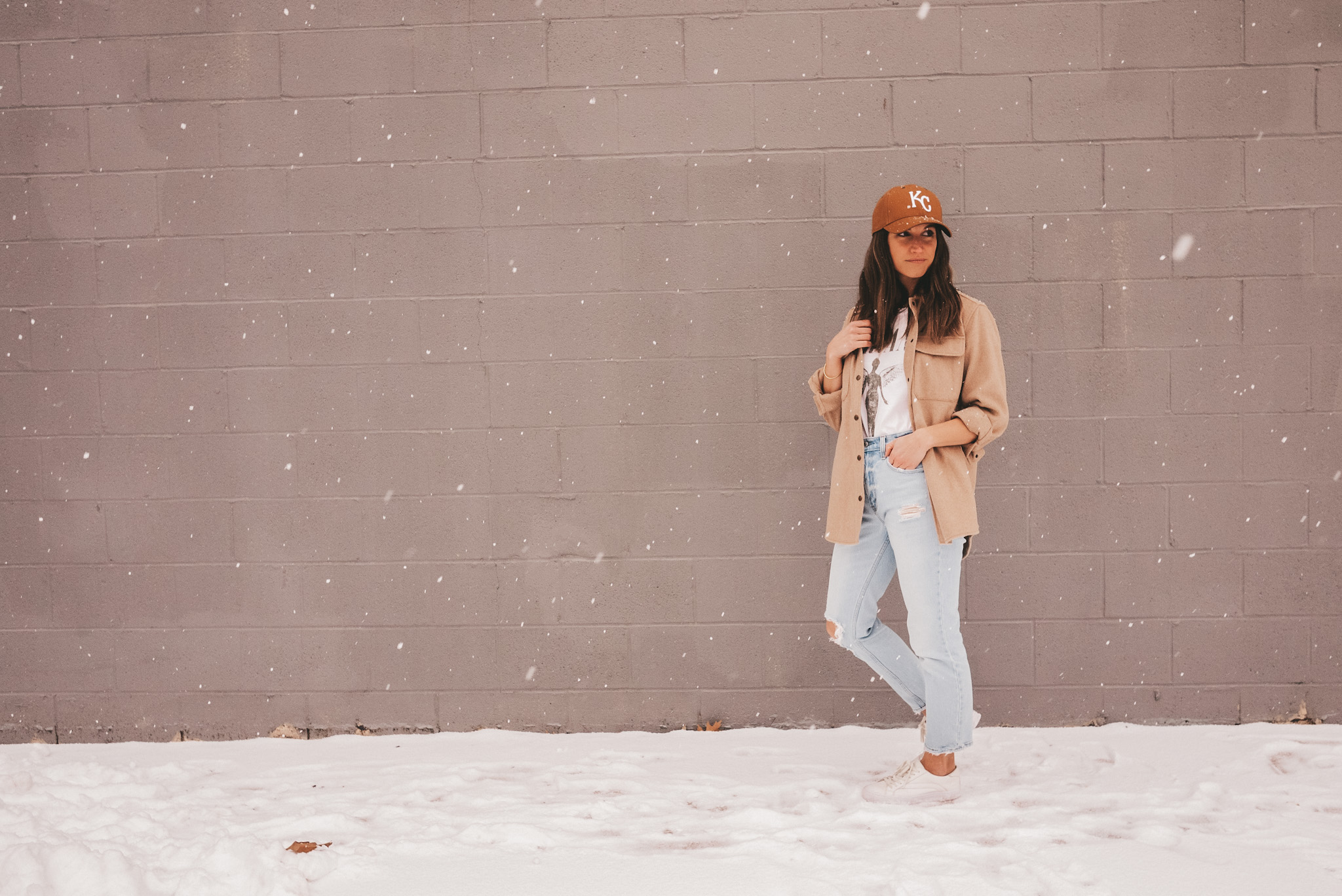 ashley standing on a snowy sidewalk with a gray brick background wearing a KC hat, tan colored shacket, white graphic tee, light washed distressed mom jeans and white sneakers