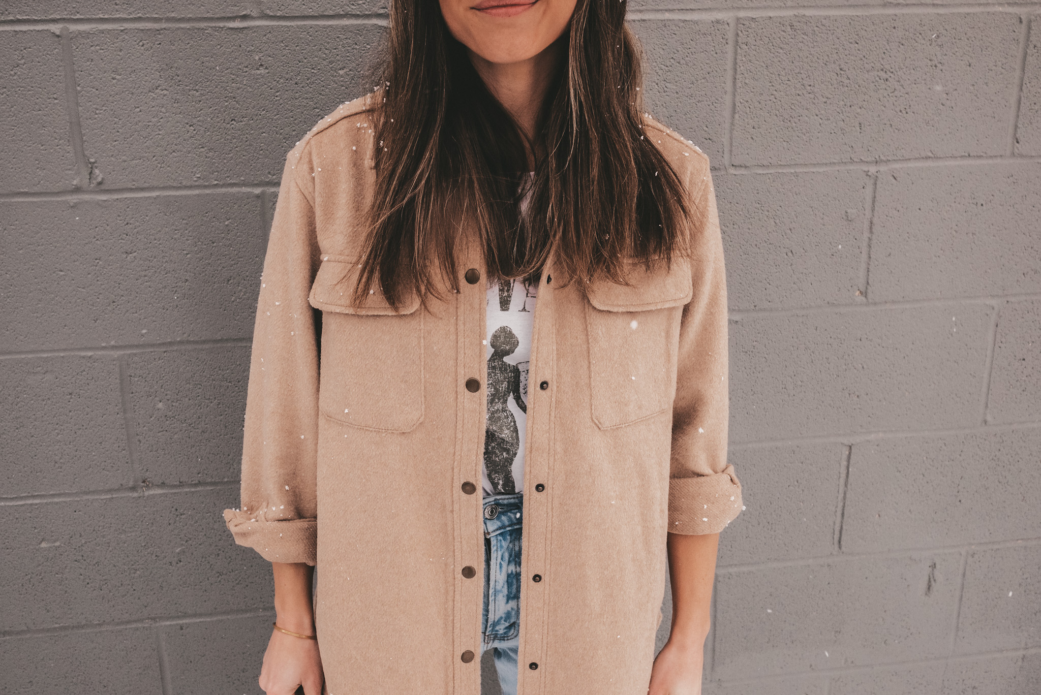 ashley wearing a tan colored shacket, a white graphic tee, and light wash jeans, standing in front of a gray brick wall