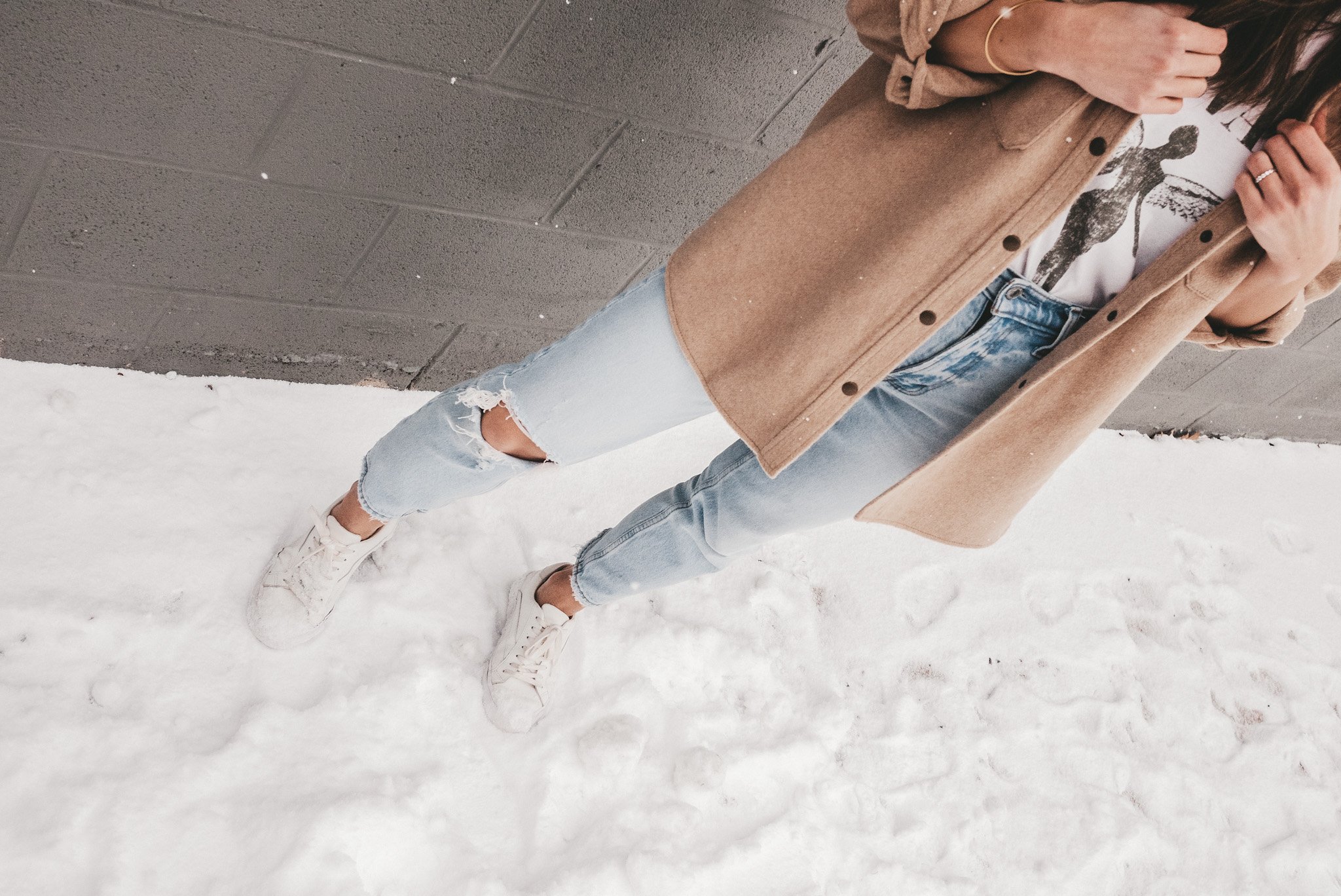 ashley standing on a snowy sidewalk with a gray brick background, wearing light wash distressed jeans, a tan colored shacket, and a white graphic tee