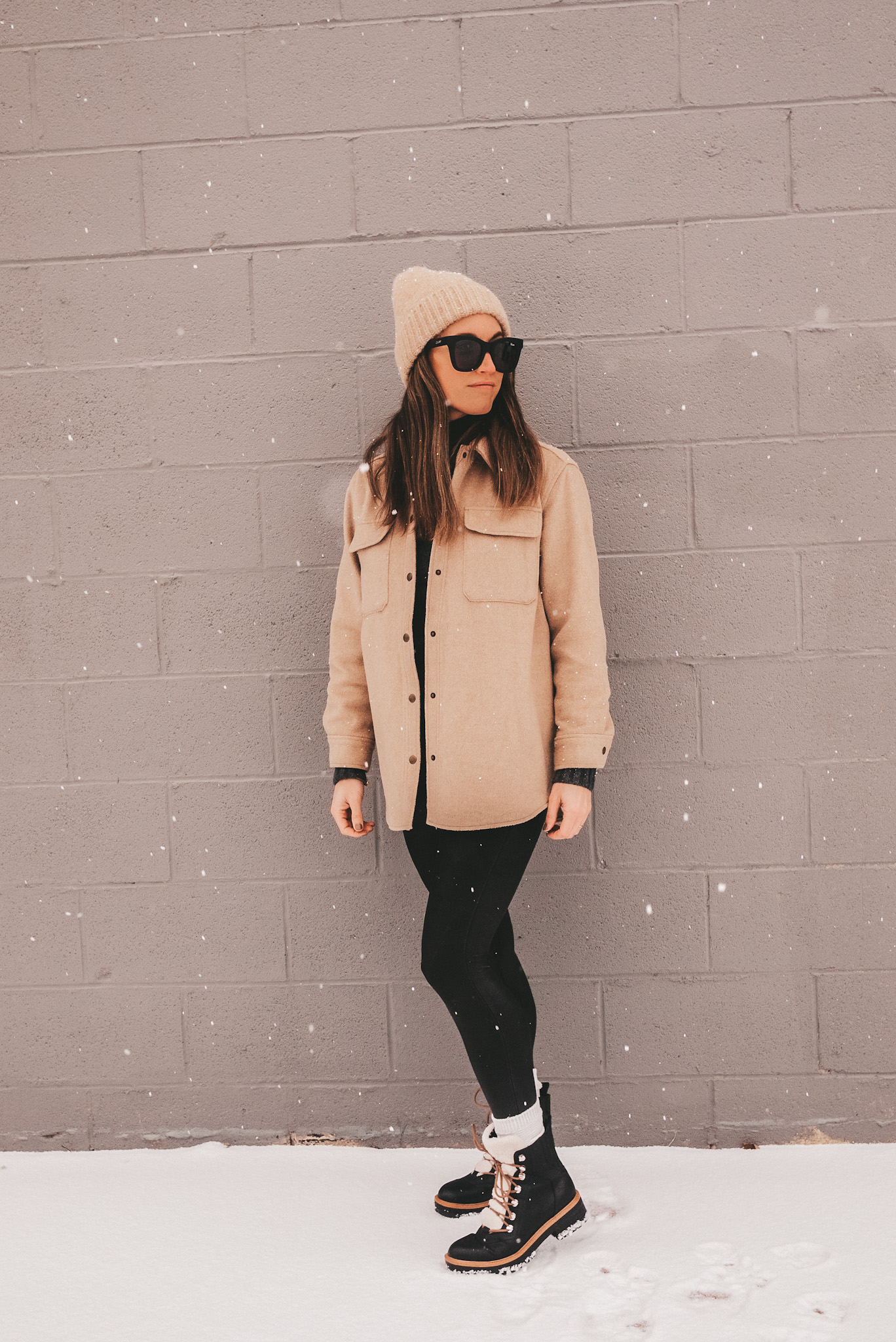Ashley wearing a tan colored shacket from Old Navy, a gray sweater from J. Crew, a tan beanie, black square frame sunglasses, Spanx faux leather leggings, black winter boots with faux sherpa lining standing in the snow with a gray brick background