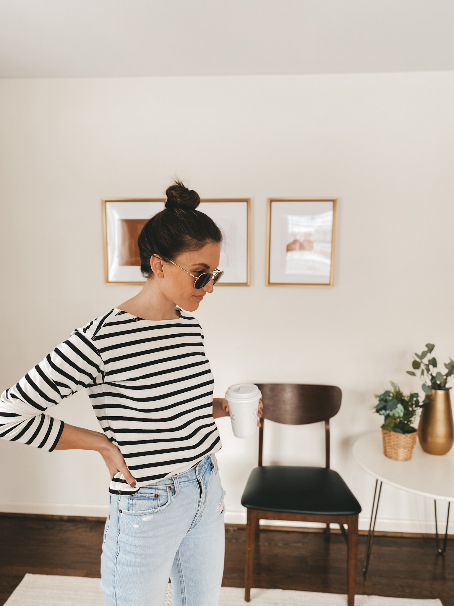 ashley wearing a striped top, aviator sunglasses, light wash distressed mom jeans from Abercrombie. She is holding a to-go coffee cup, there is a chair and 2 plants