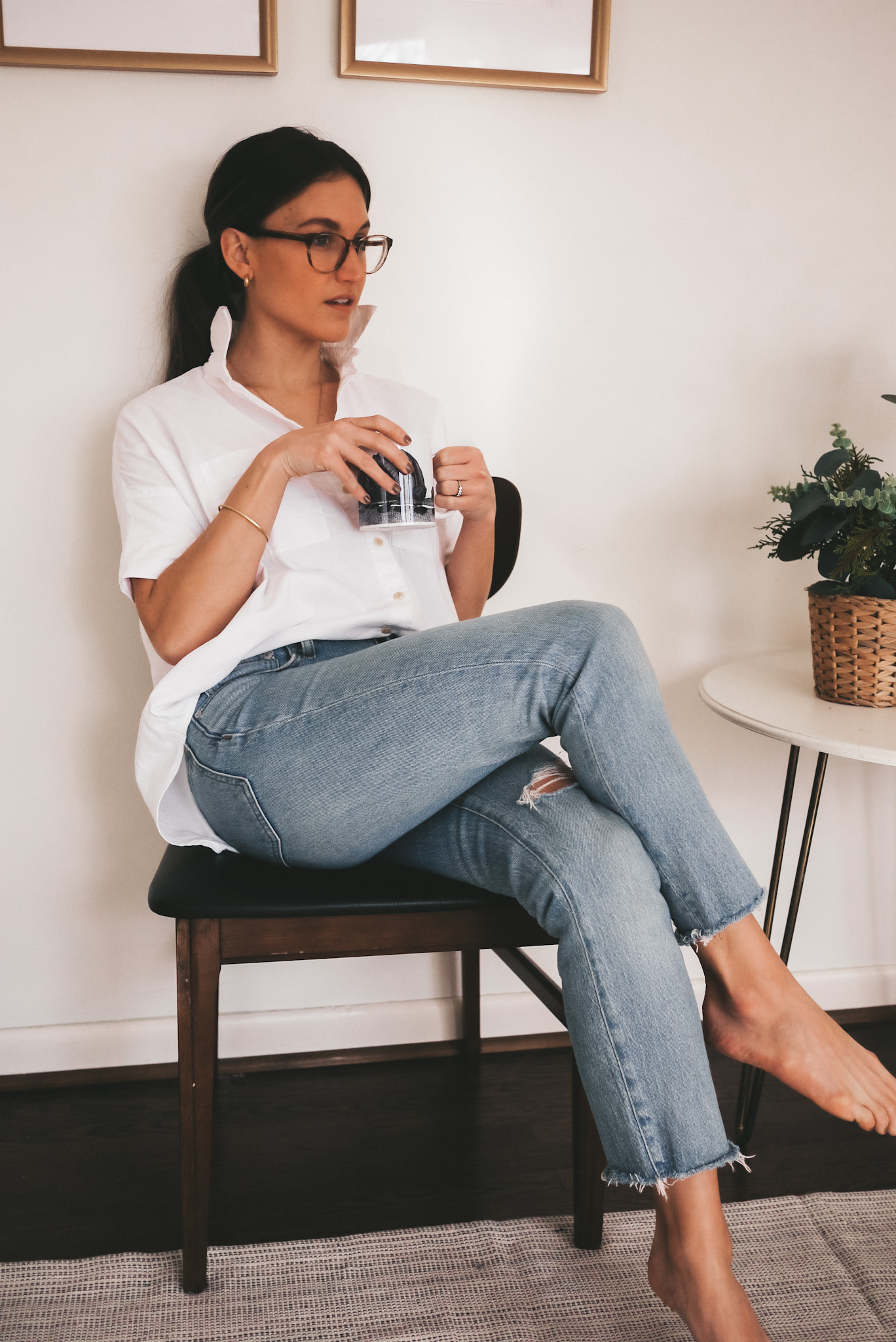 ashley wearing a Madewell white button down shirt and light wash distressed jeans sitting in a chair and holding a coffee mug