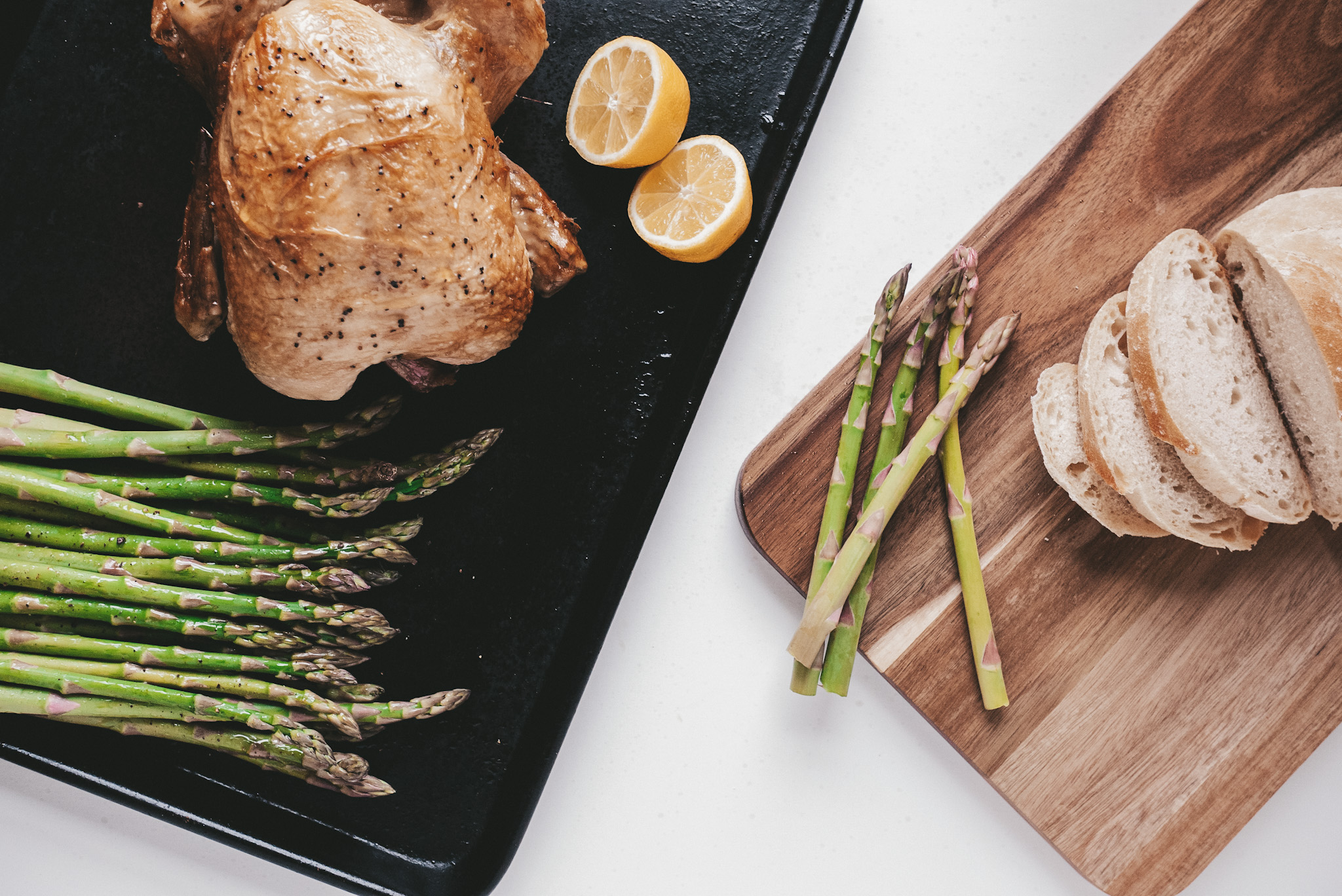 rotisserie chicken and asparagus on a baking sheet, a wooden cutting board with sliced bread on a white background