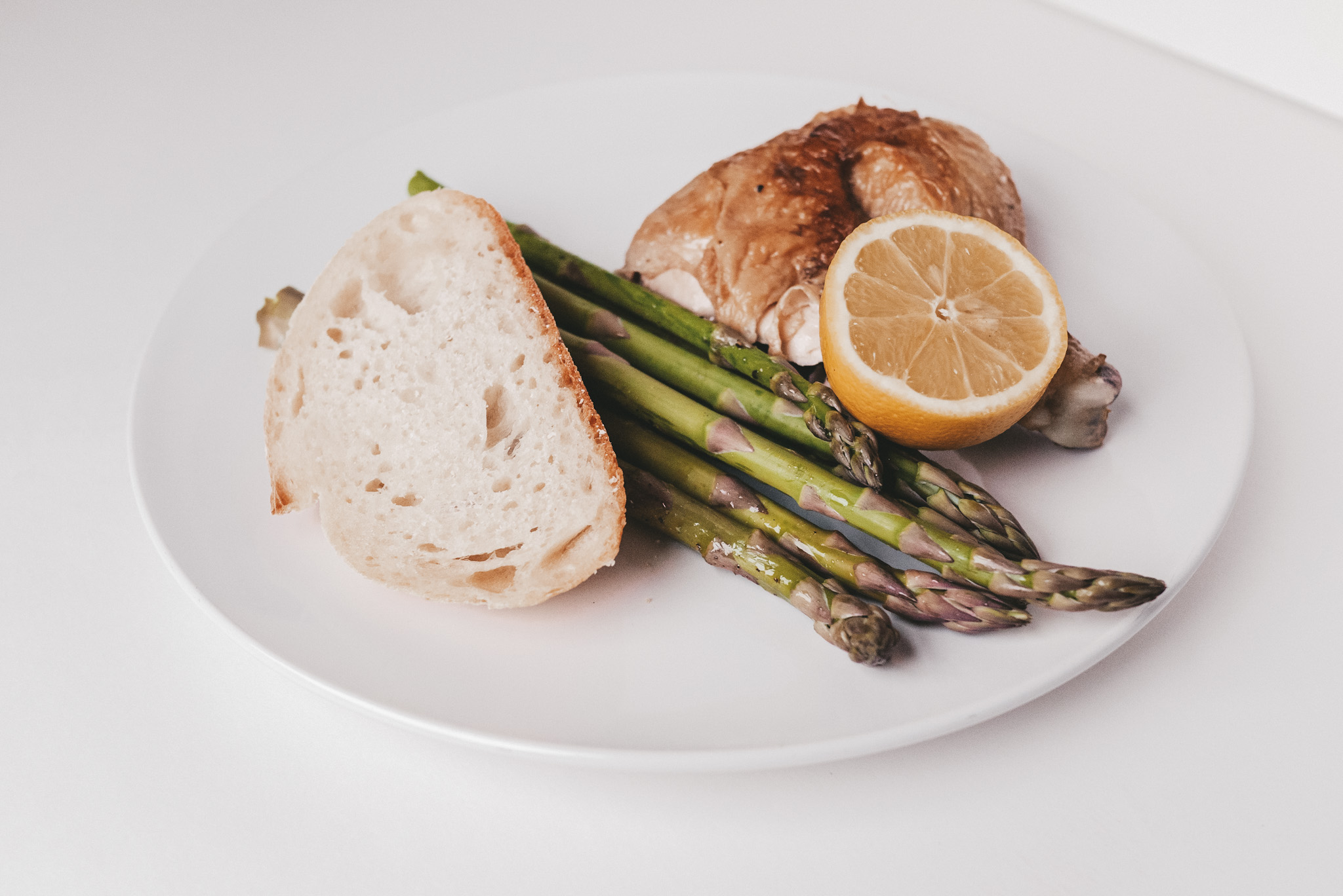 a white plate with a slice of bread, asparagus, chicken thigh and drumstick, and a lemon sliced