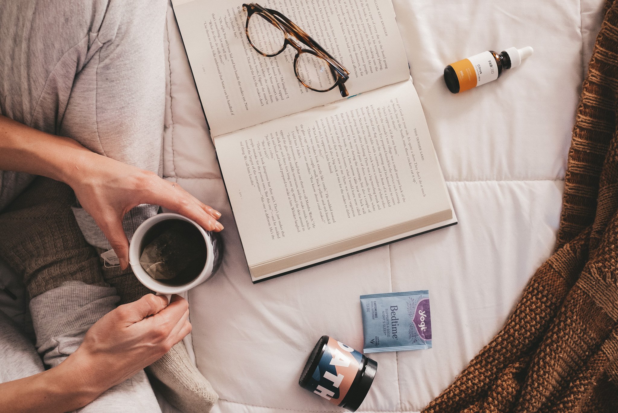 objects to improve your sleep; a white bedspread with a girl in gray sweatpants and taupe socks holding a mug of tea; a book, body lotion, package of tea, glasses and CBD bottle are on the bedspread
