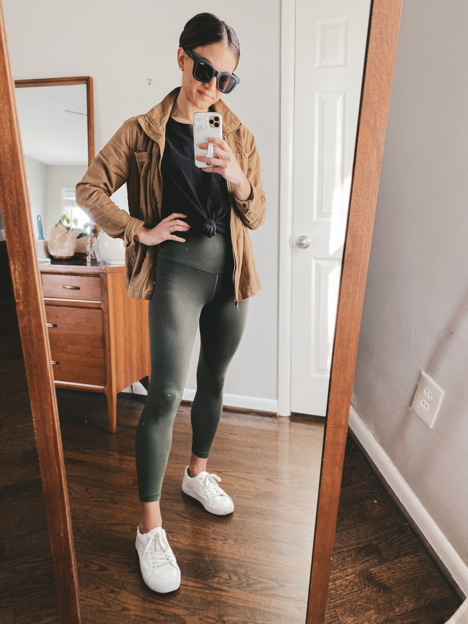 ashley wearing an Old Navy tan linen utility jacket, a black Everlane Tee tied at the waist, olive green leggings from Aerie, white sneakers from Madewell and black square frame sunglasses from Quay