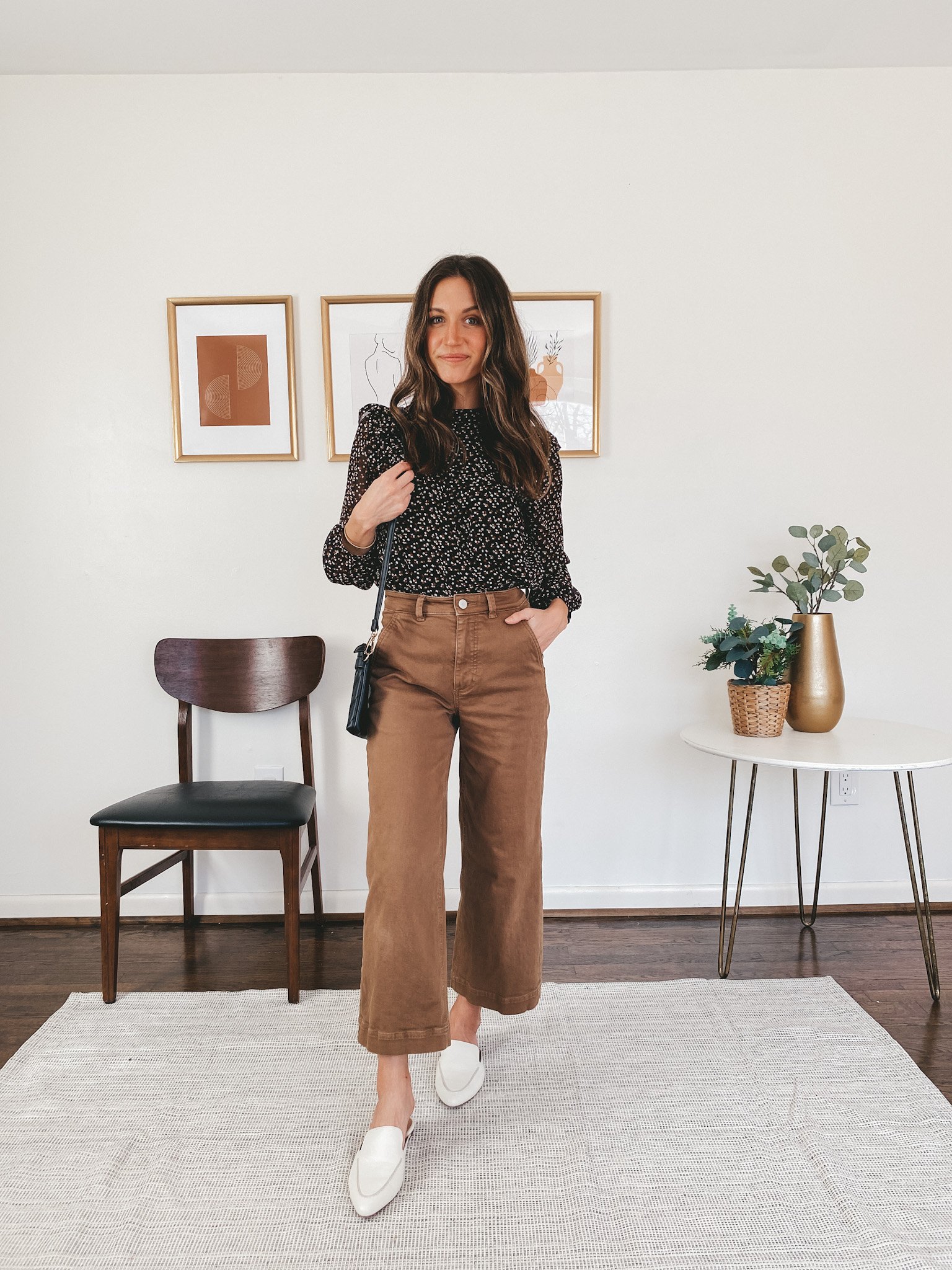 ashley wearing a Madewell ruffle puff sleeve top in black with a small flower pattern, Everlane wide leg pants in ochre, Madewell white mules and a black quilted crossbody bag