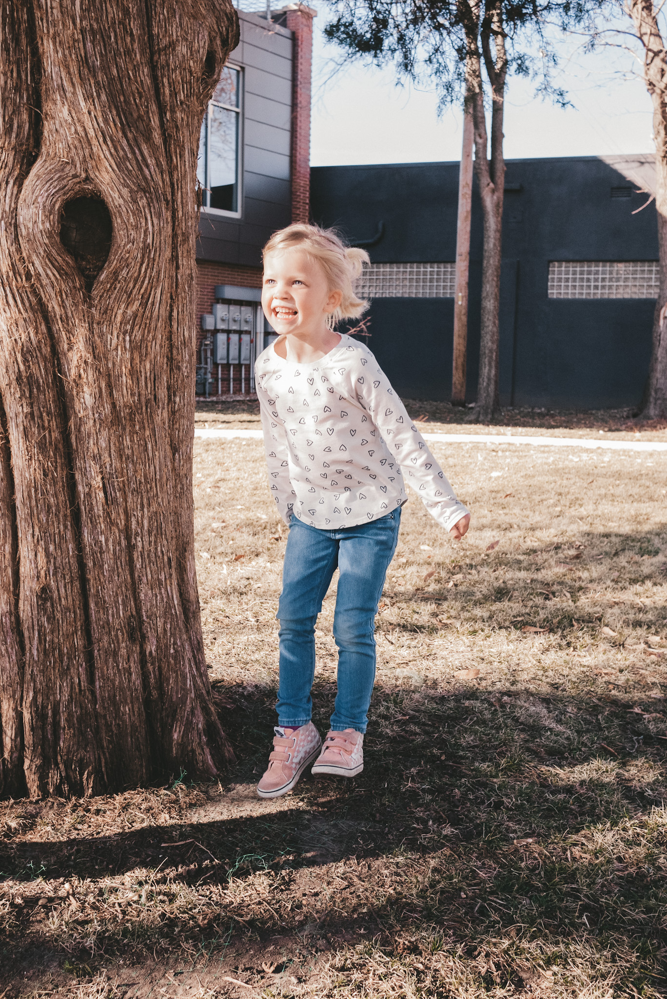 a little girl wearing a heart print long sleeve tee, jeans and pink high top Vans, jumping next to a tree