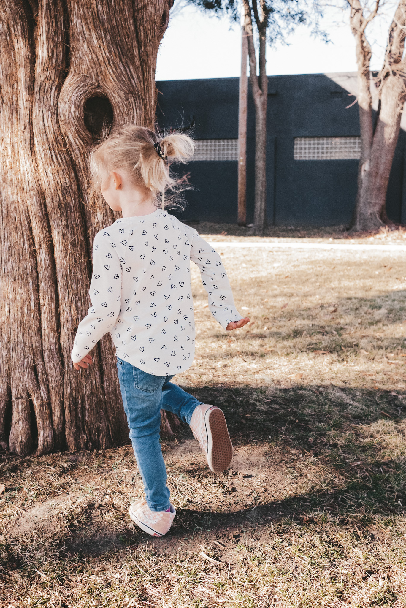 a little girl wearing a heart print long sleeve tee, jeans and pink high top Vans, standing next to a tree