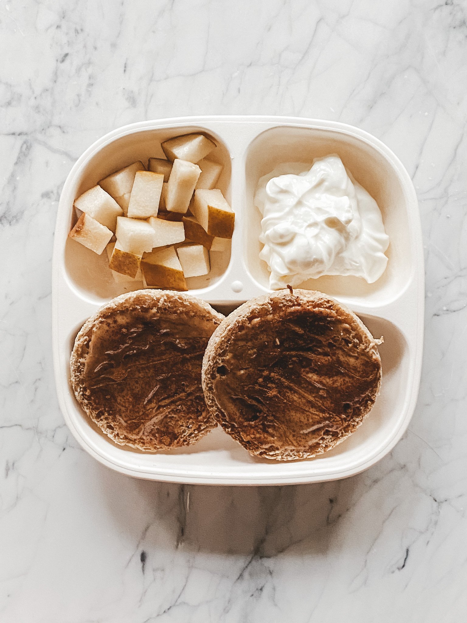 a white toddler plate with a toasted english muffins with peanut butter, honey and cinnamon, diced pears, and greek yogurt