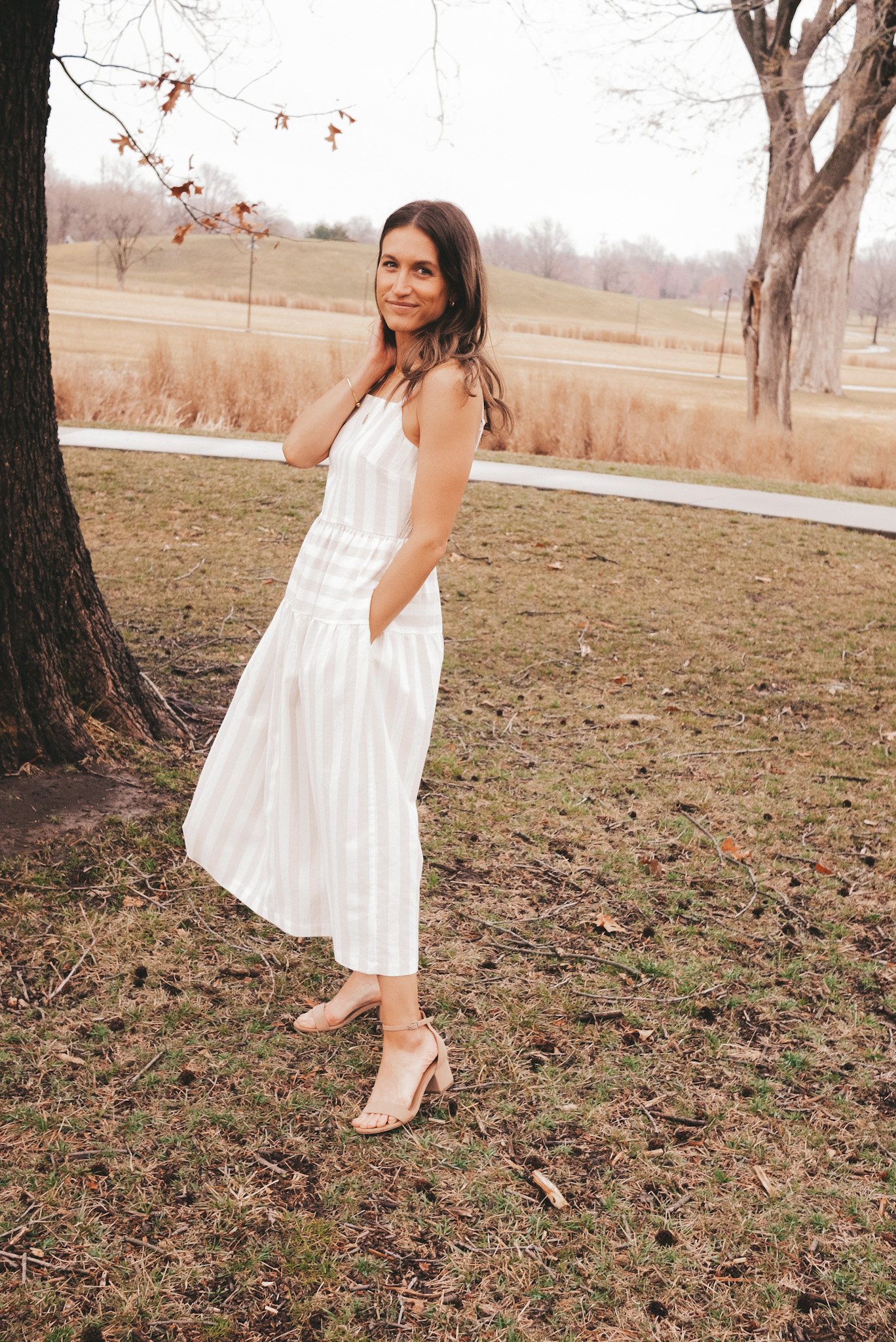 ashley wearing a white and tan striped midi dress with block heeled sandals standing in a park