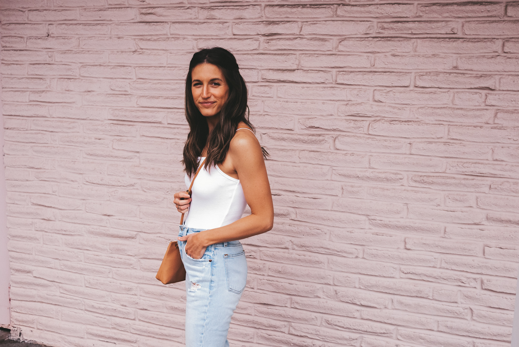 ashley wearing a white spaghetti strap bodysuit, and light wash jeans, with a camel colored crossbody bag standing in front of a gray brick wall