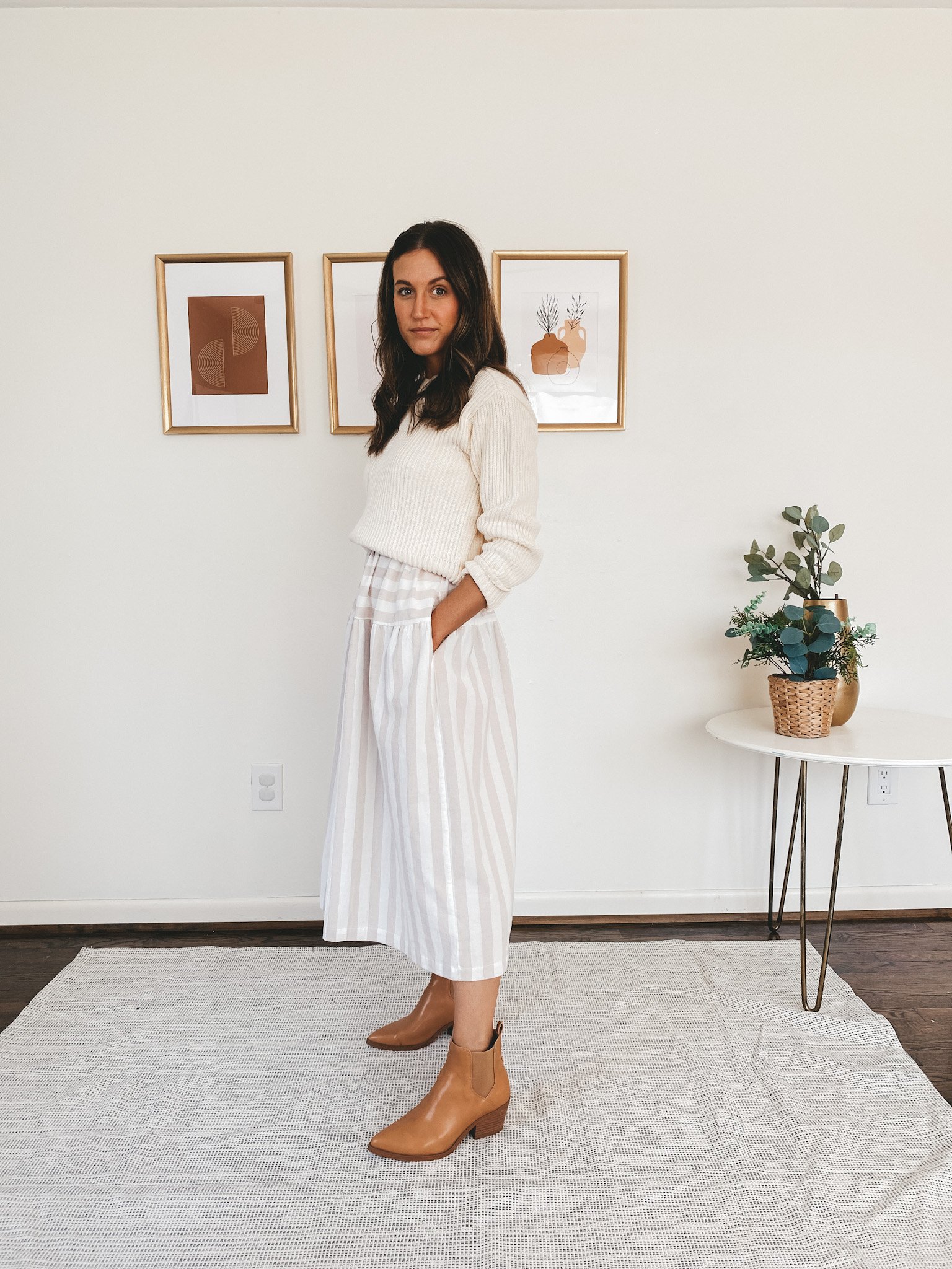 ashley wearing a tan and white striped midi dress with a cream sweater over it, and cognac ankle boots