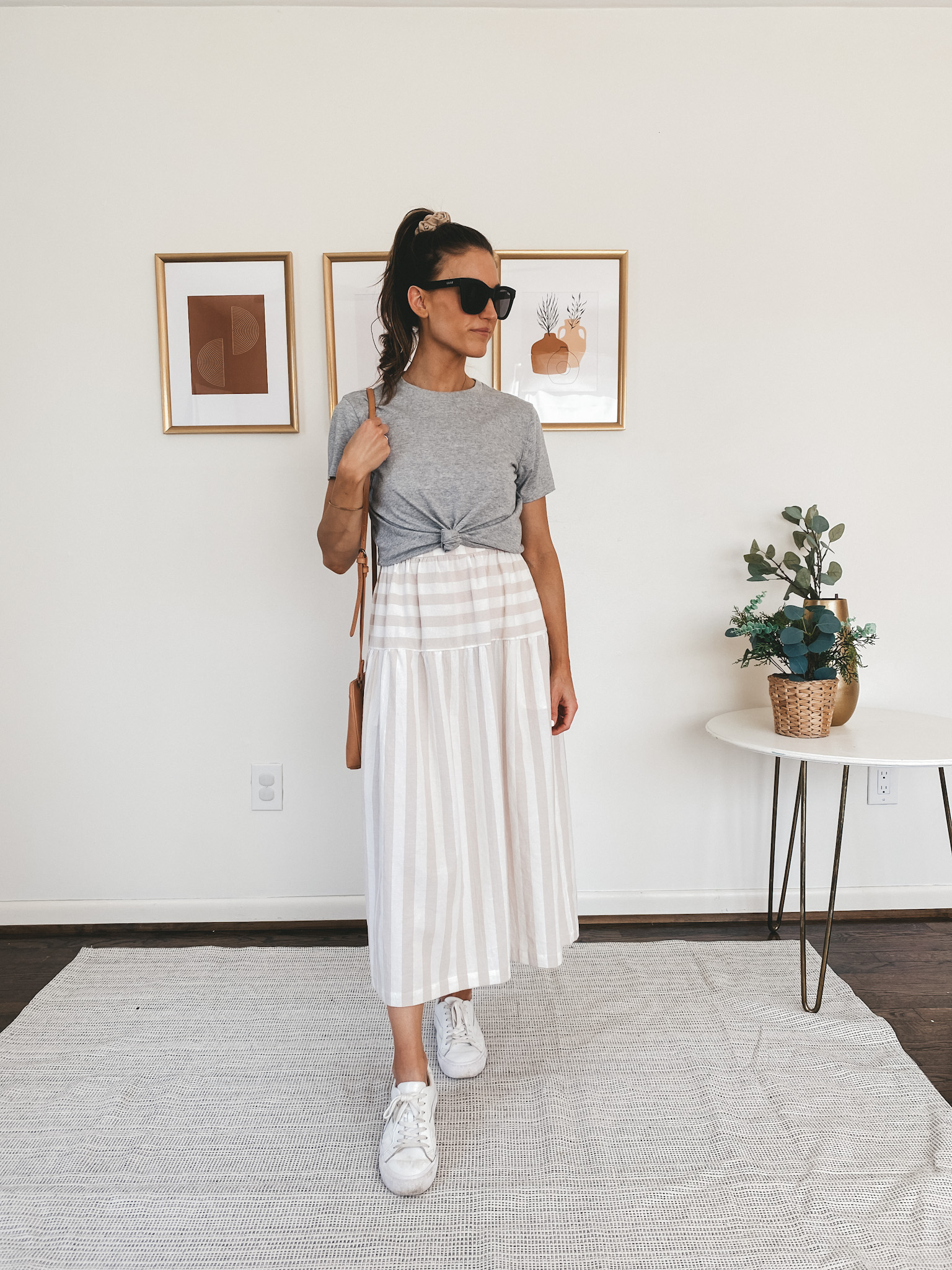 ashley wearing an Everlane gray tee over a tan and white striped midi dress, Quay Australia Sunglasses, Madewell white sneakers, and a Target tan crossbody bag