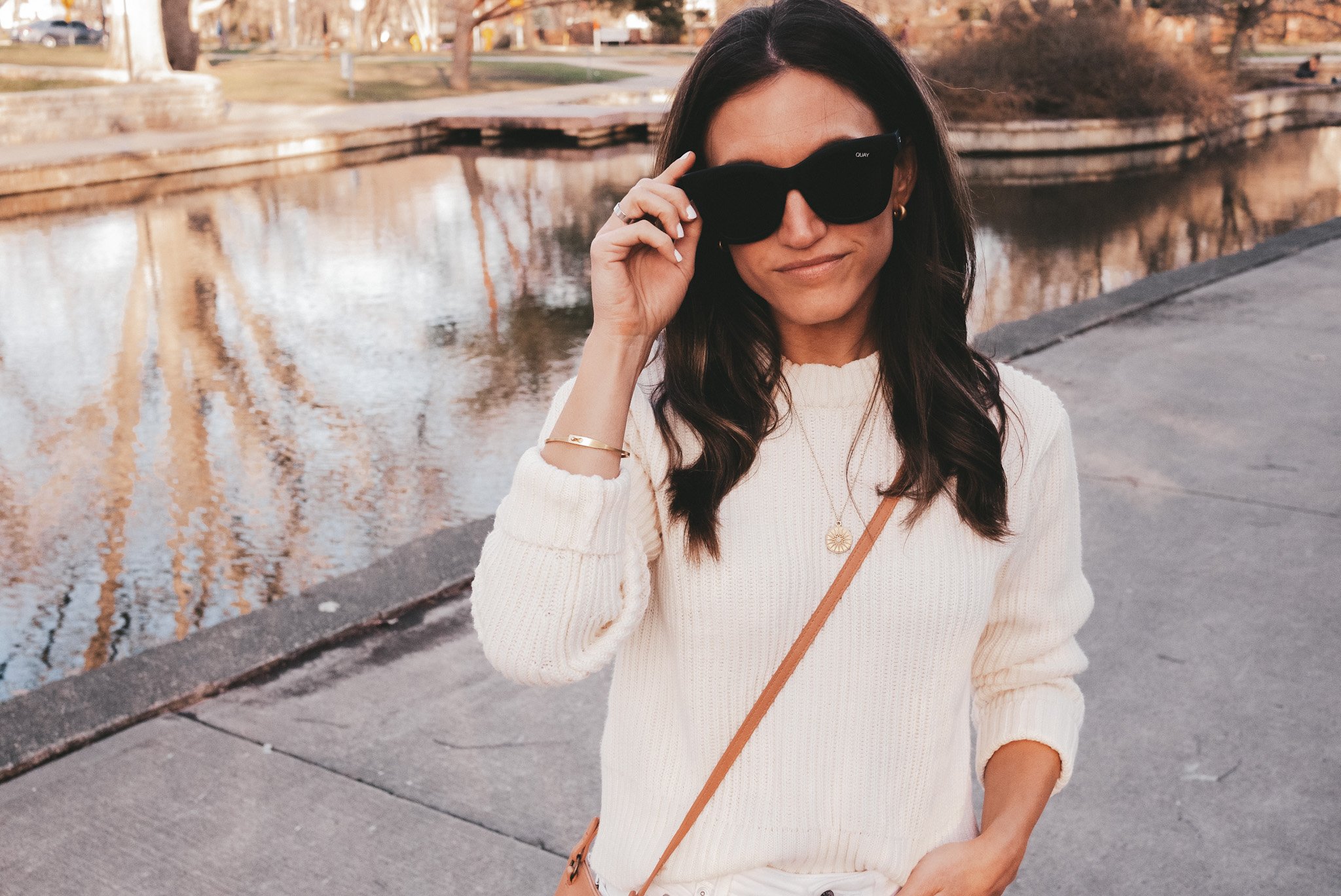 ashley standing by a pond wearing a cream colored sweater and sunglasses