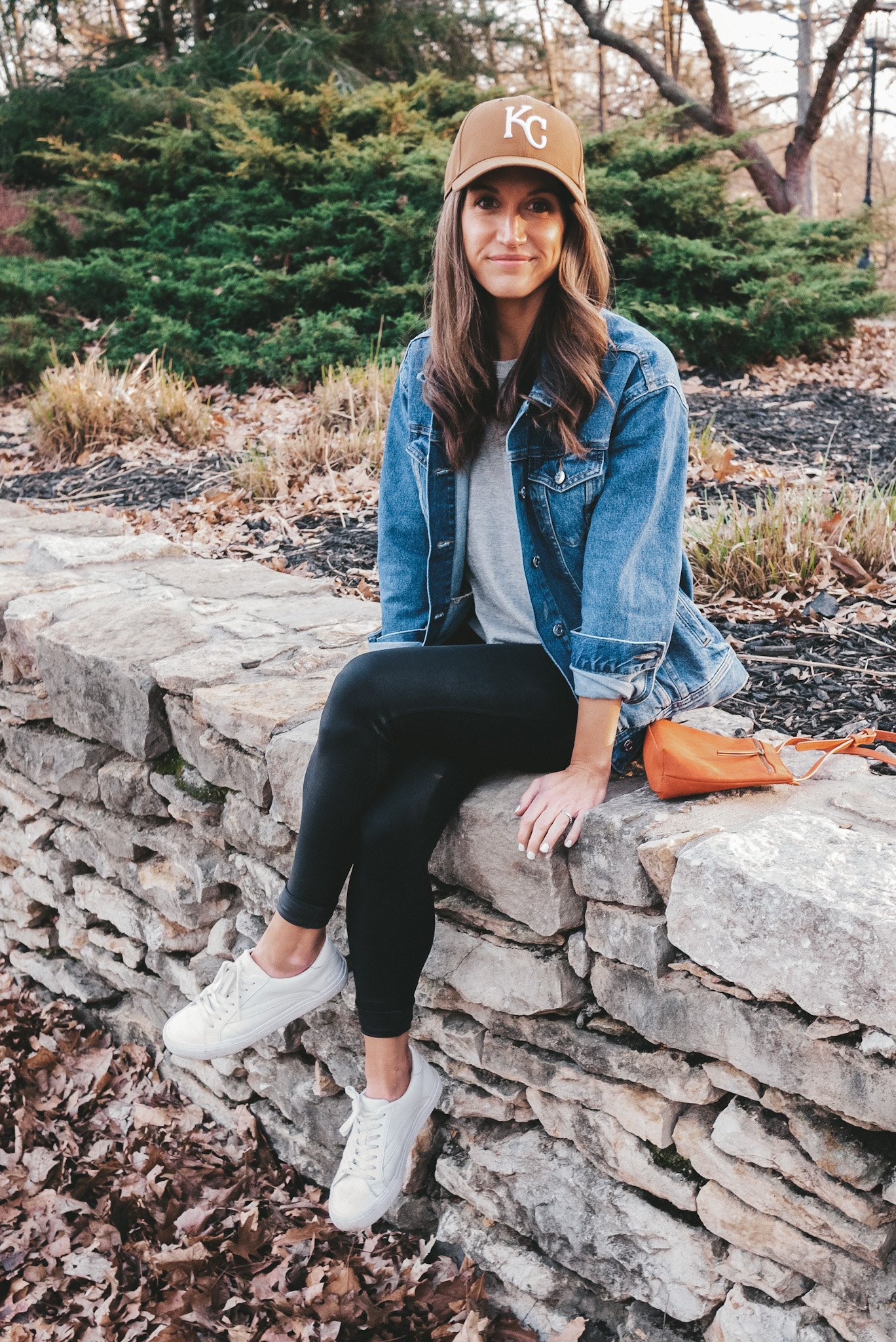 ashley sitting on a stone wall in a park wearing a denim jacket, gray tee, brown KC baseball hat, faux leather leggings and white sneakers