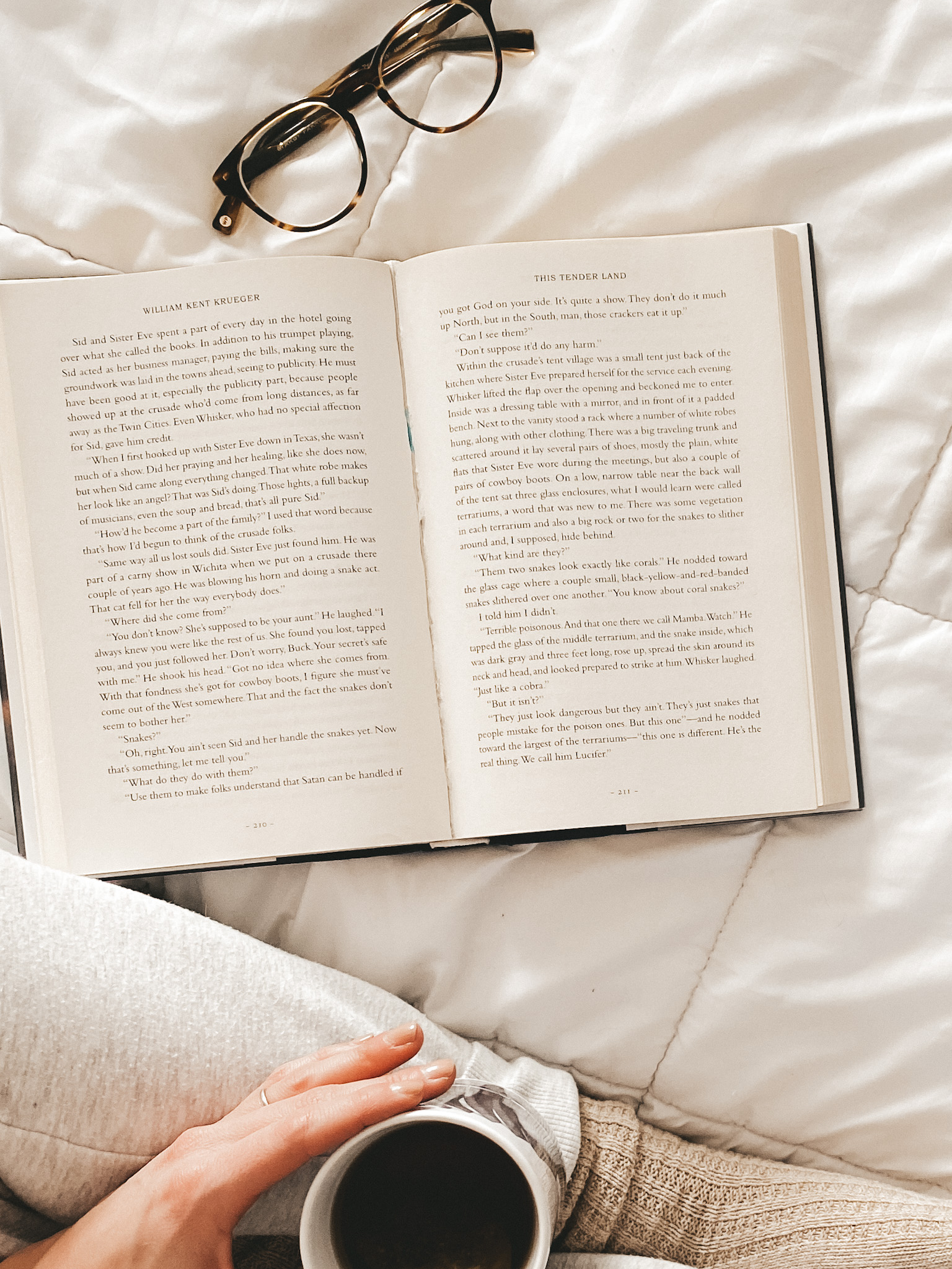 a book open on a white comforter, a pair of glasses, a woman