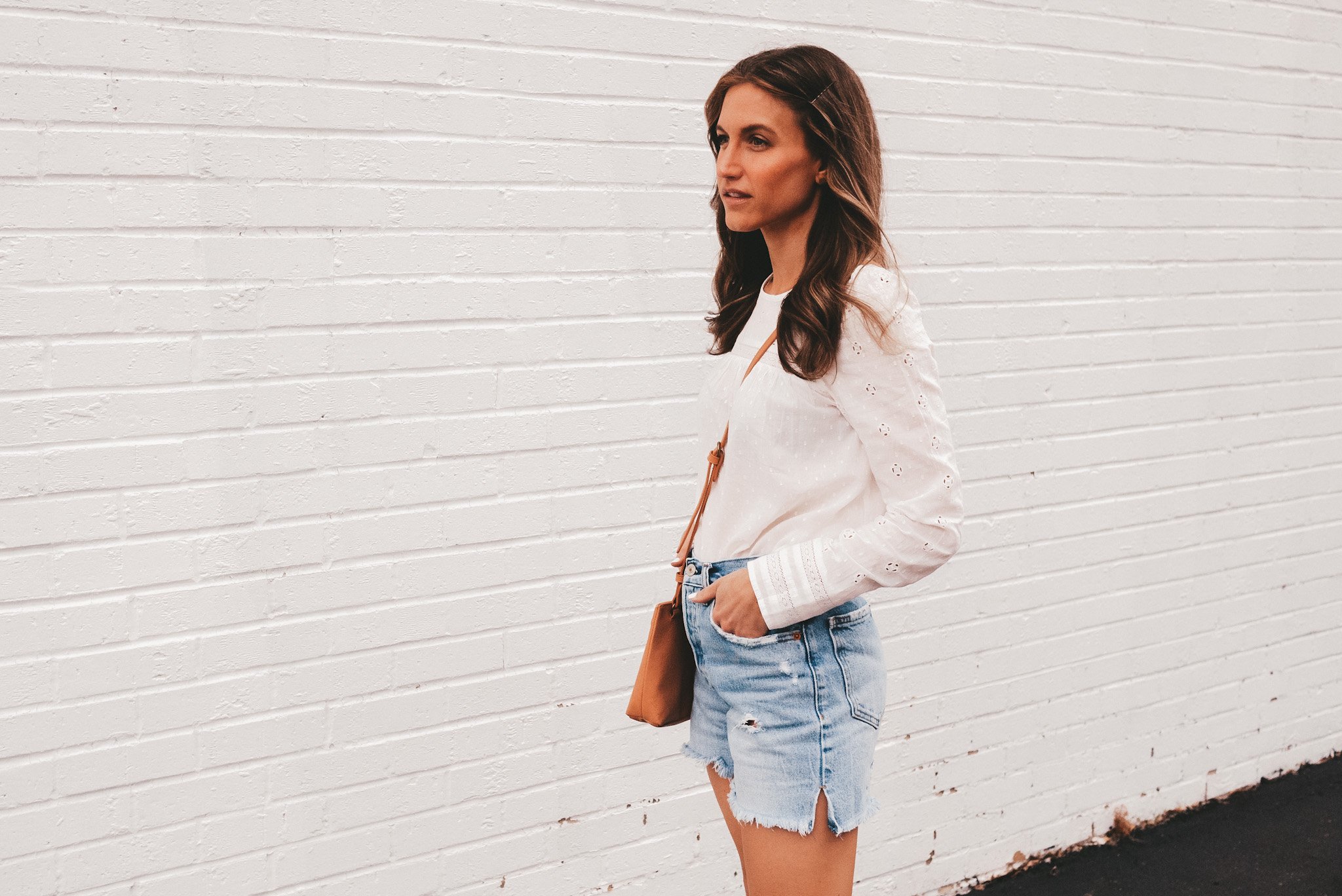 ashley wearing a white eyelet top and cut off denim shorts standing in front of a white brick wall