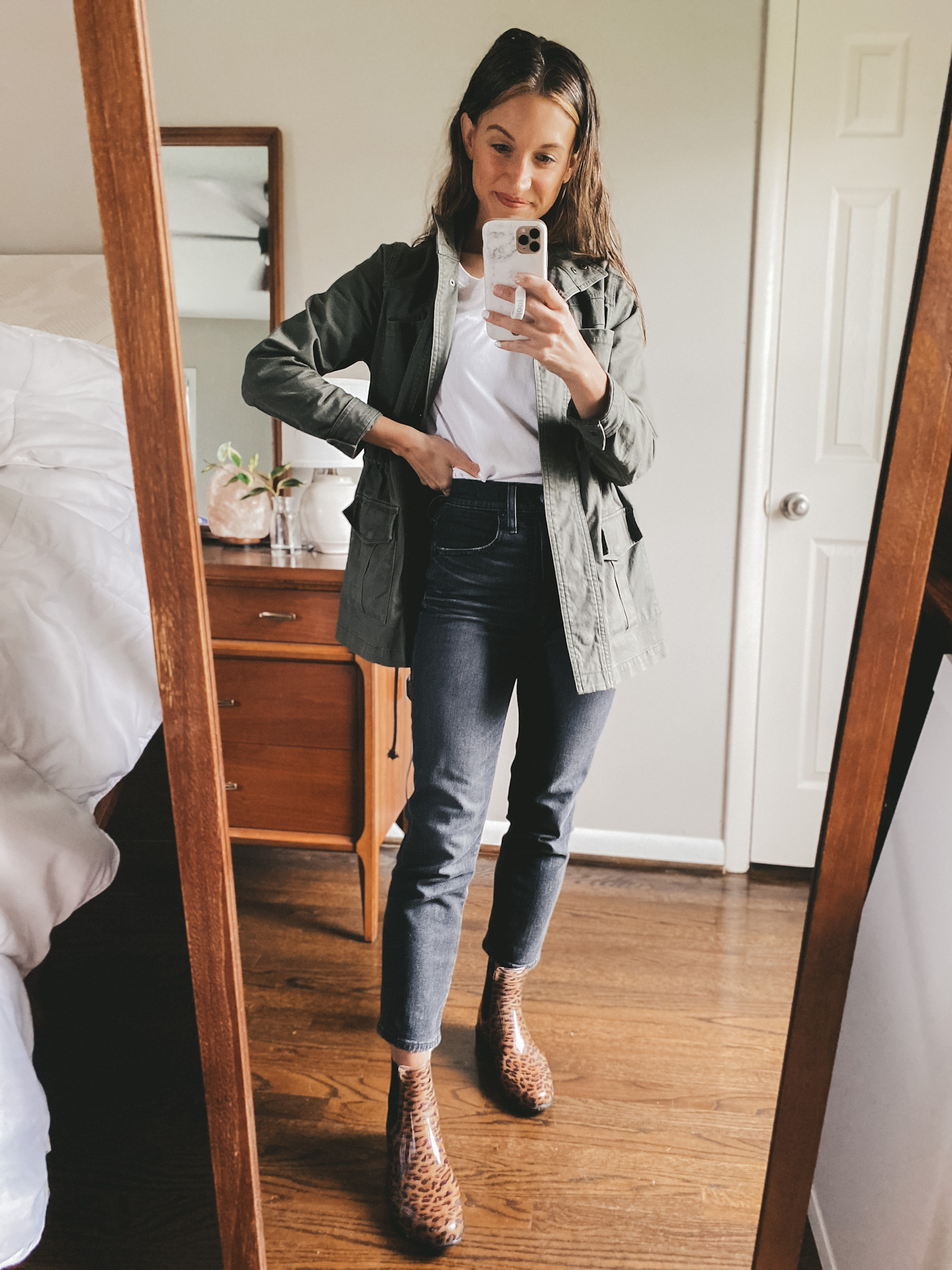 ashley wearing a white tee, green utility jacket, washed black straight leg jeans, and leopard print rain boots
