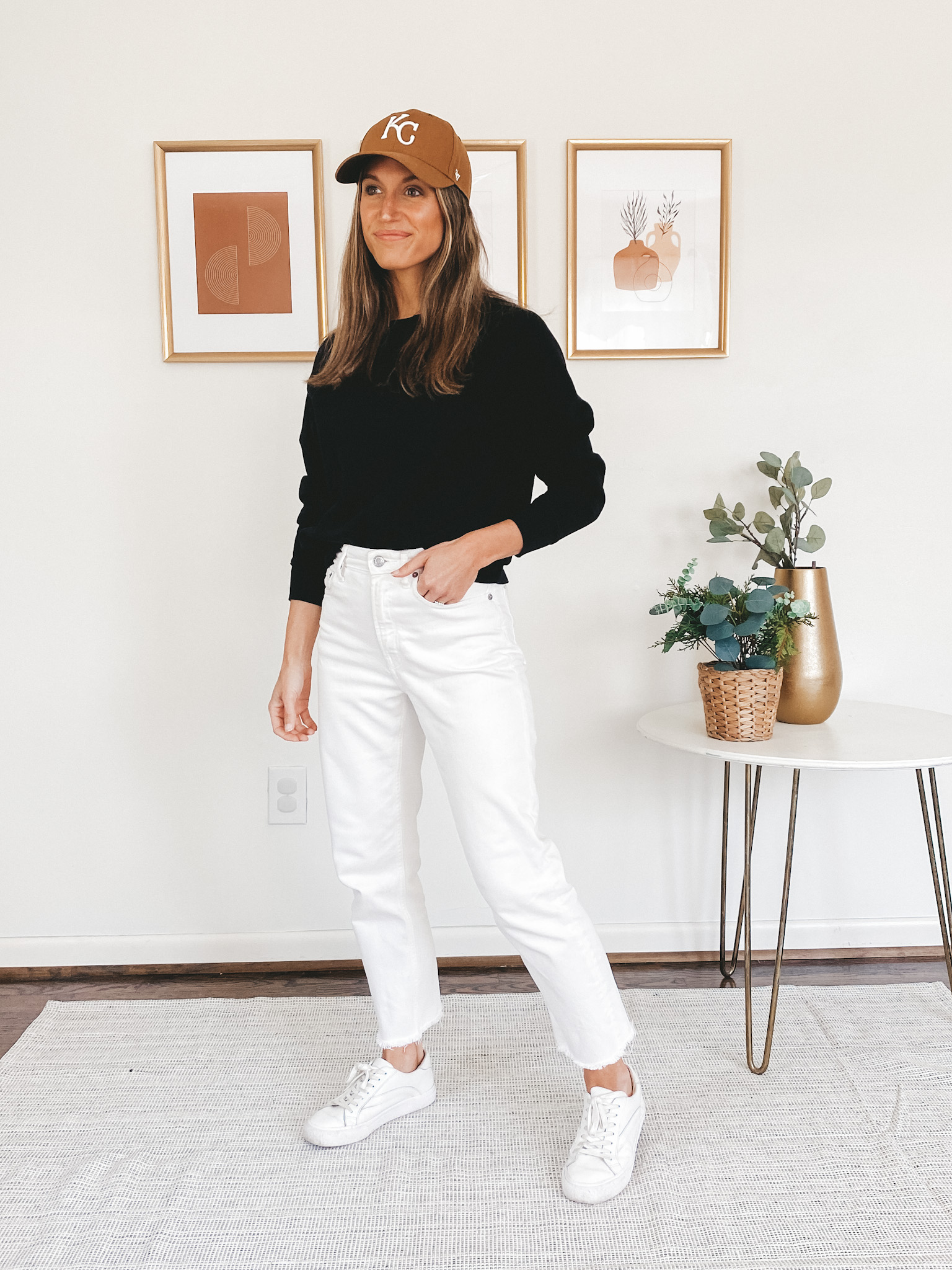 ashley wearing a black crewneck sweatshirt, white denim, white sneakers, and a KC baseball cap
