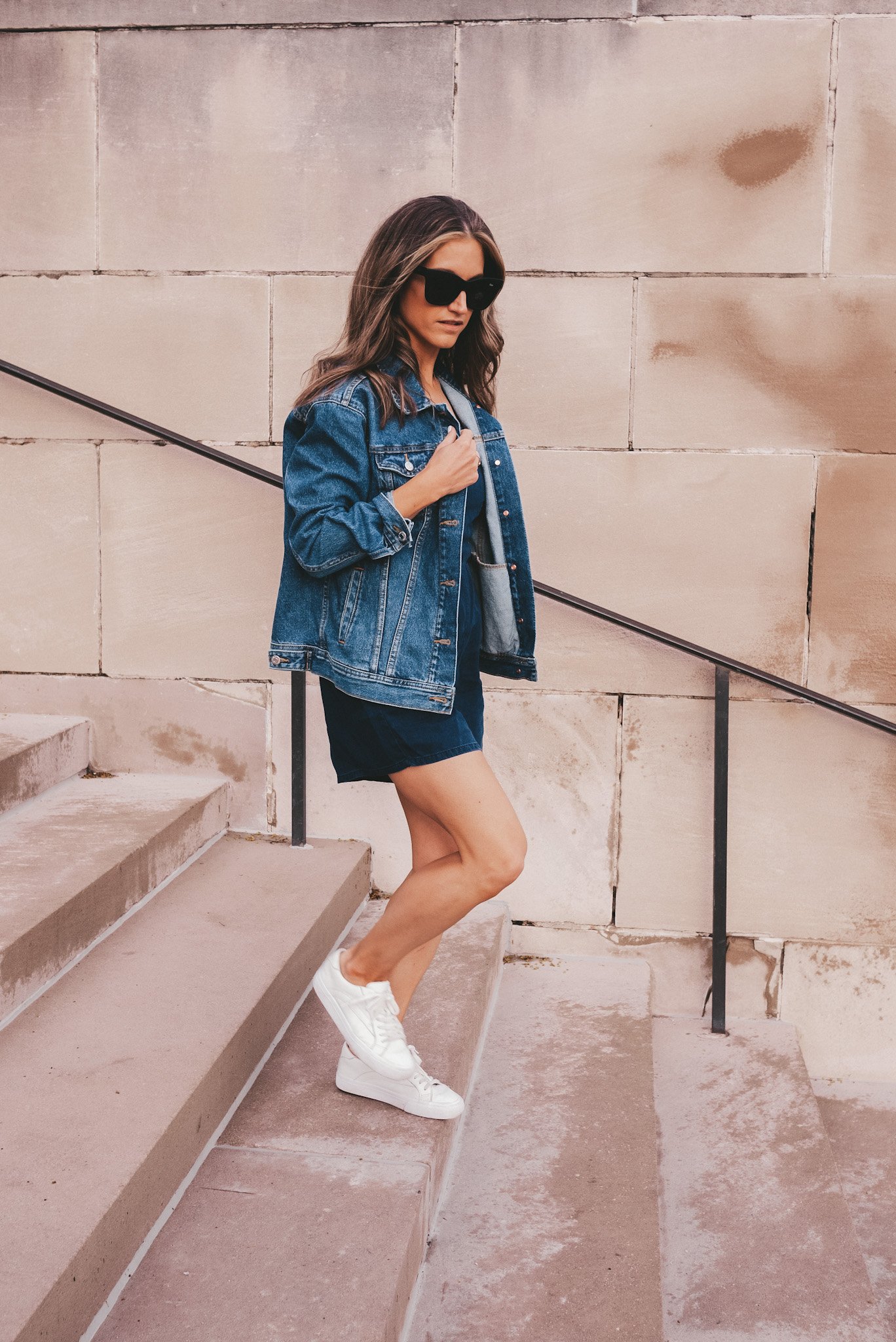 ashley wearing a chambray romper, oversized denim jacket, white sneakers, and square frame black sunglasses walking down stairs at The Nelson Atkins Museum in Kansas City