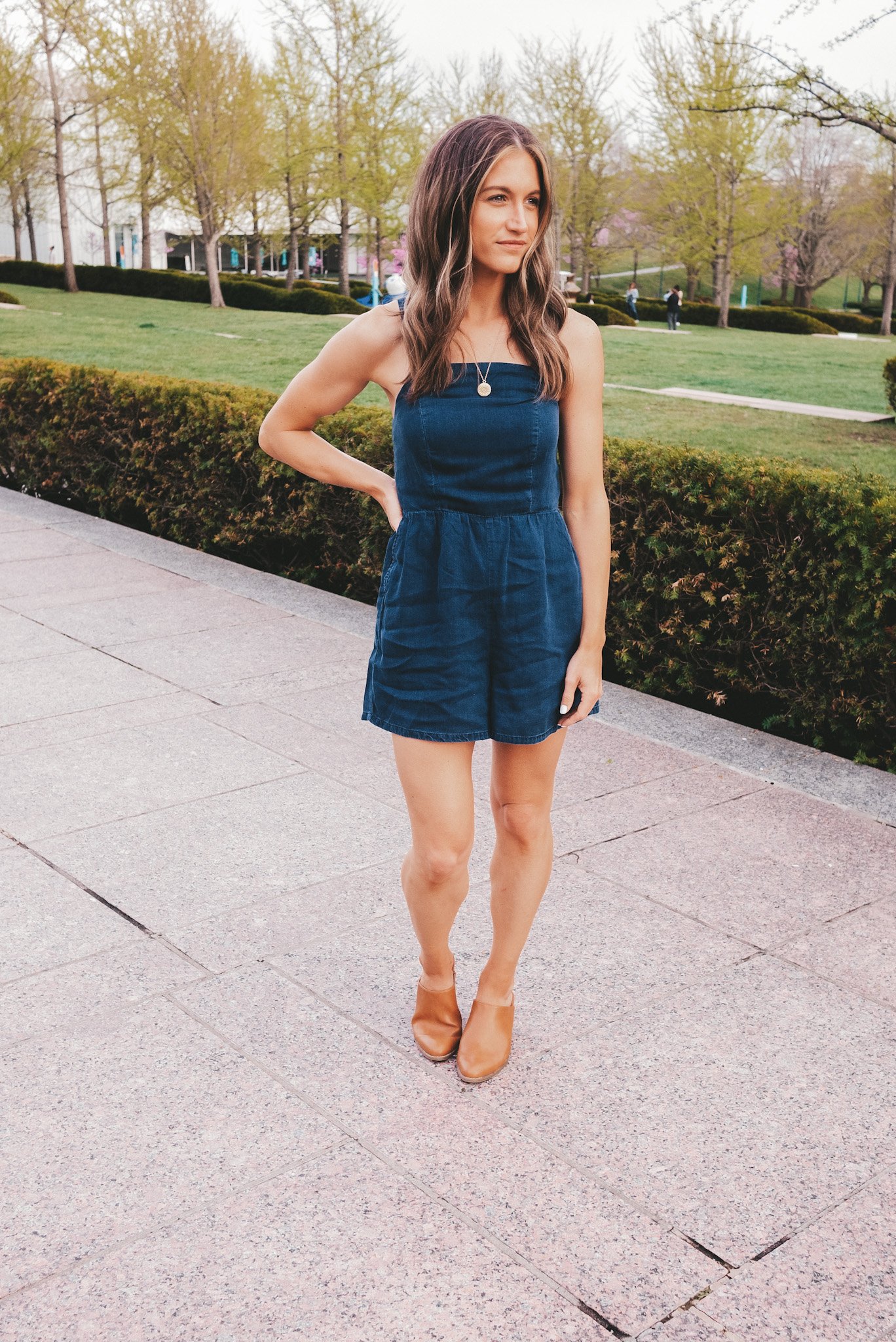 ashley wearing a chambray romper and heeled mules standing next to a hedge at The Nelson-Atkins Museum in Kansas City