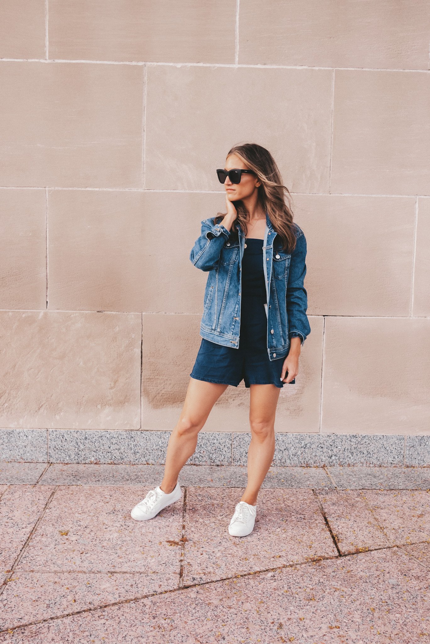 Ashley wearing a chambray romper, oversized denim jacket, white sneakers, and black sunglasses standing in front of a beige stone wall at The Nelson Atkin