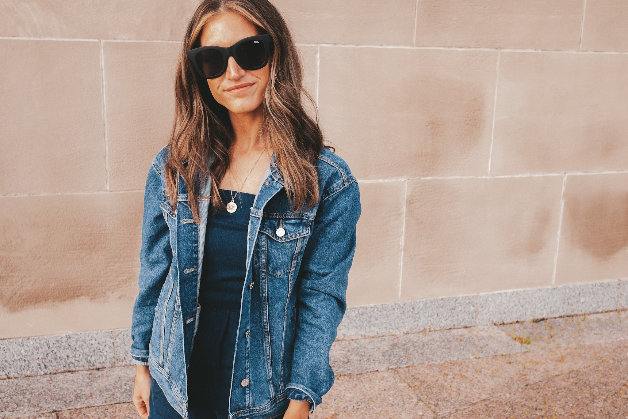 Ashley wearing a chambray romper, oversized denim jacket, white sneakers, and black sunglasses standing in front of a beige stone wall at The Nelson Atkin