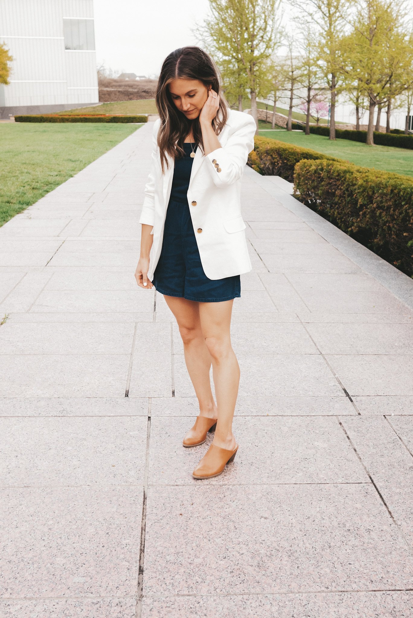 ashley wearing a chambray romper, white blazer and heeled mules standing next to a hedge at The Nelson-Atkins Museum in Kansas City