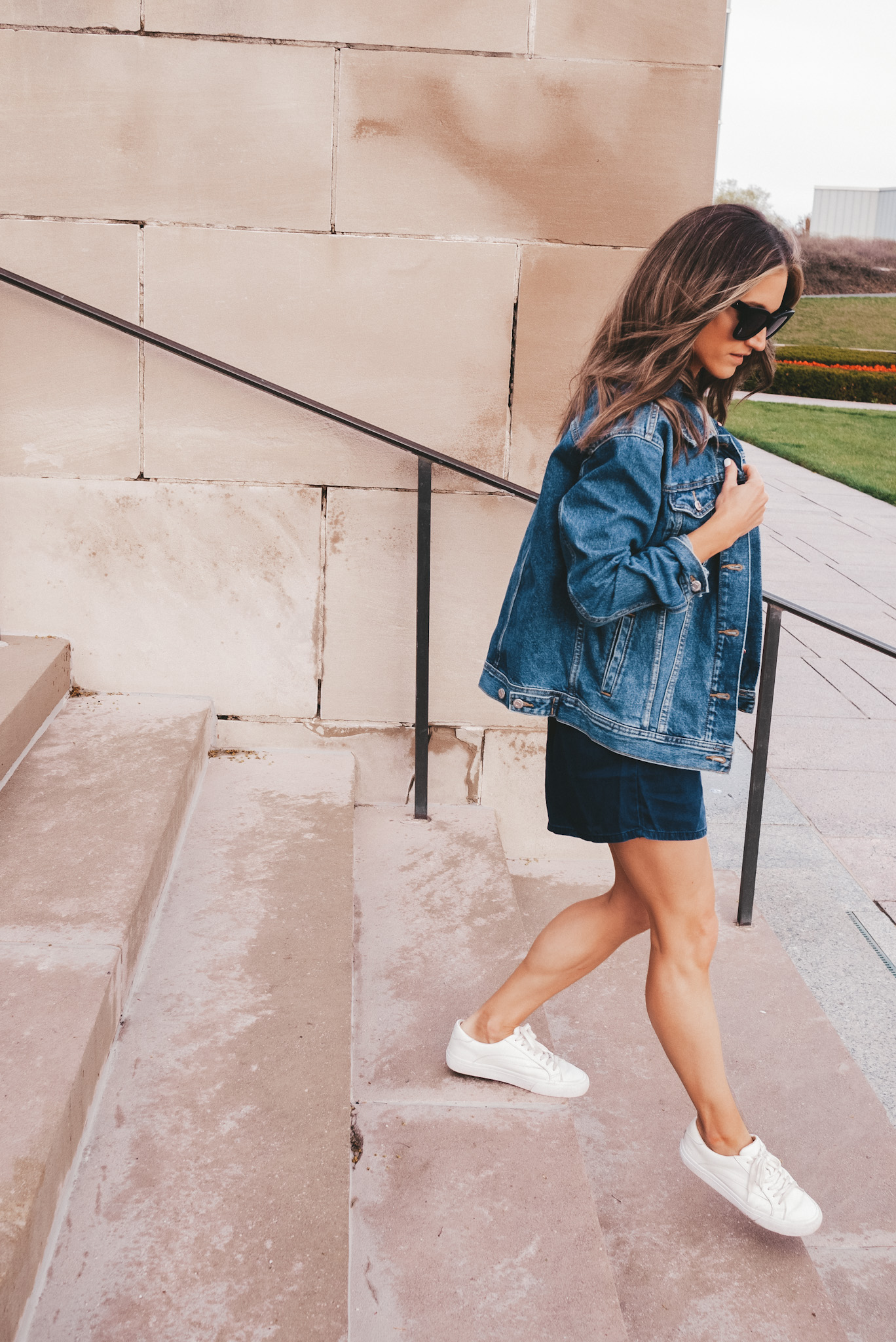 ashley wearing a chambray romper, oversized denim jacket, white sneakers, and square frame black sunglasses walking down stairs at The Nelson Atkins Museum in Kansas City