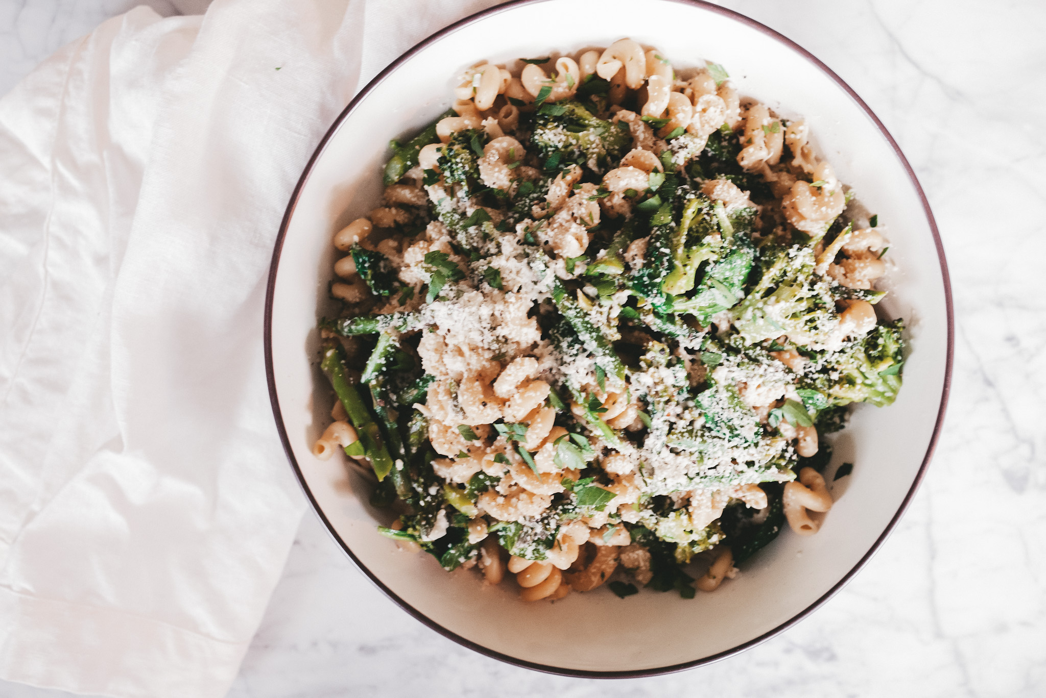 a large bowl of pasta with seasonal produce on a white marble table with a linen tea towel