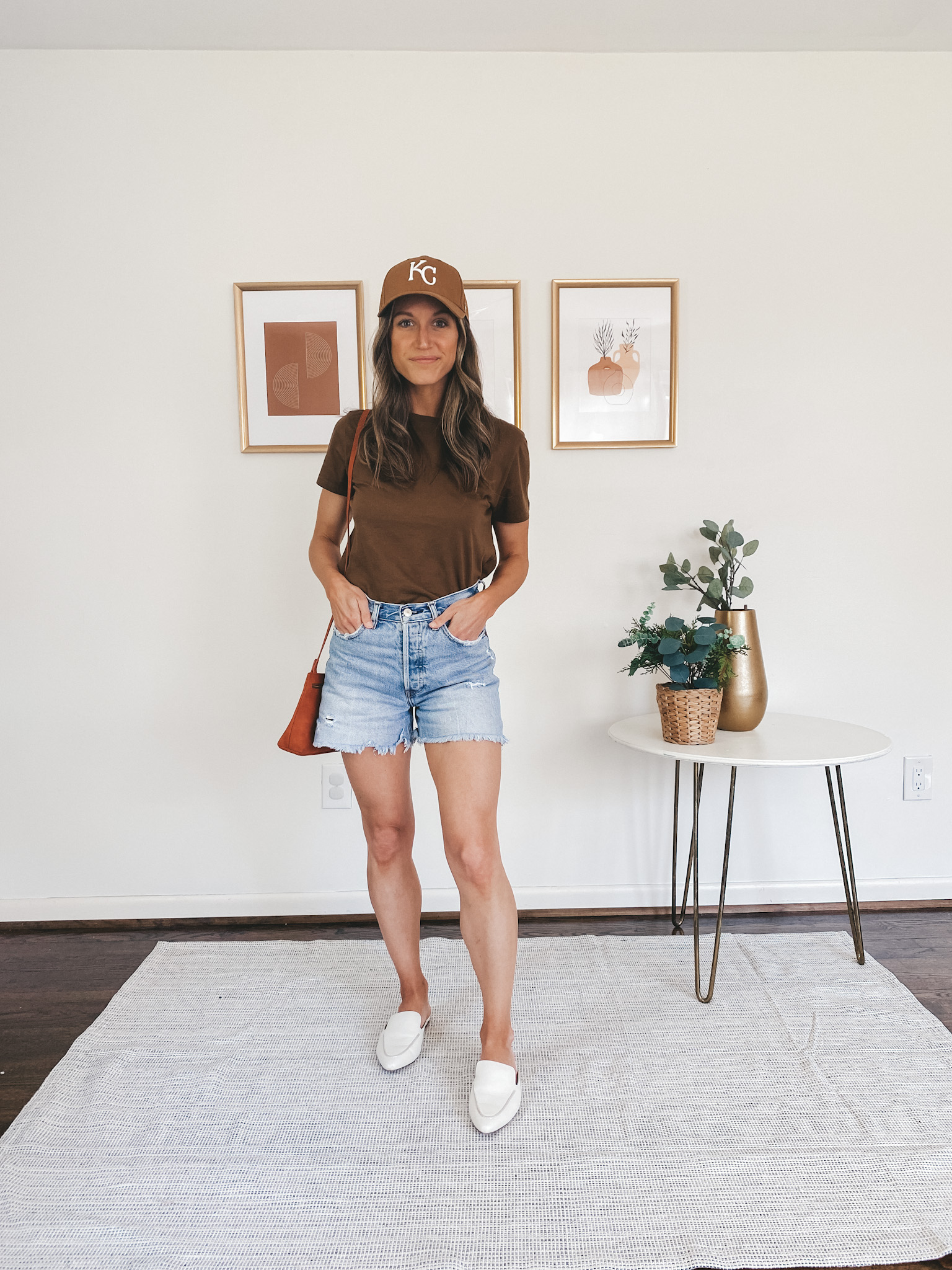 ashley wearing a brown tee, denim cut off shorts, white mules and a brown KC baseball cap
