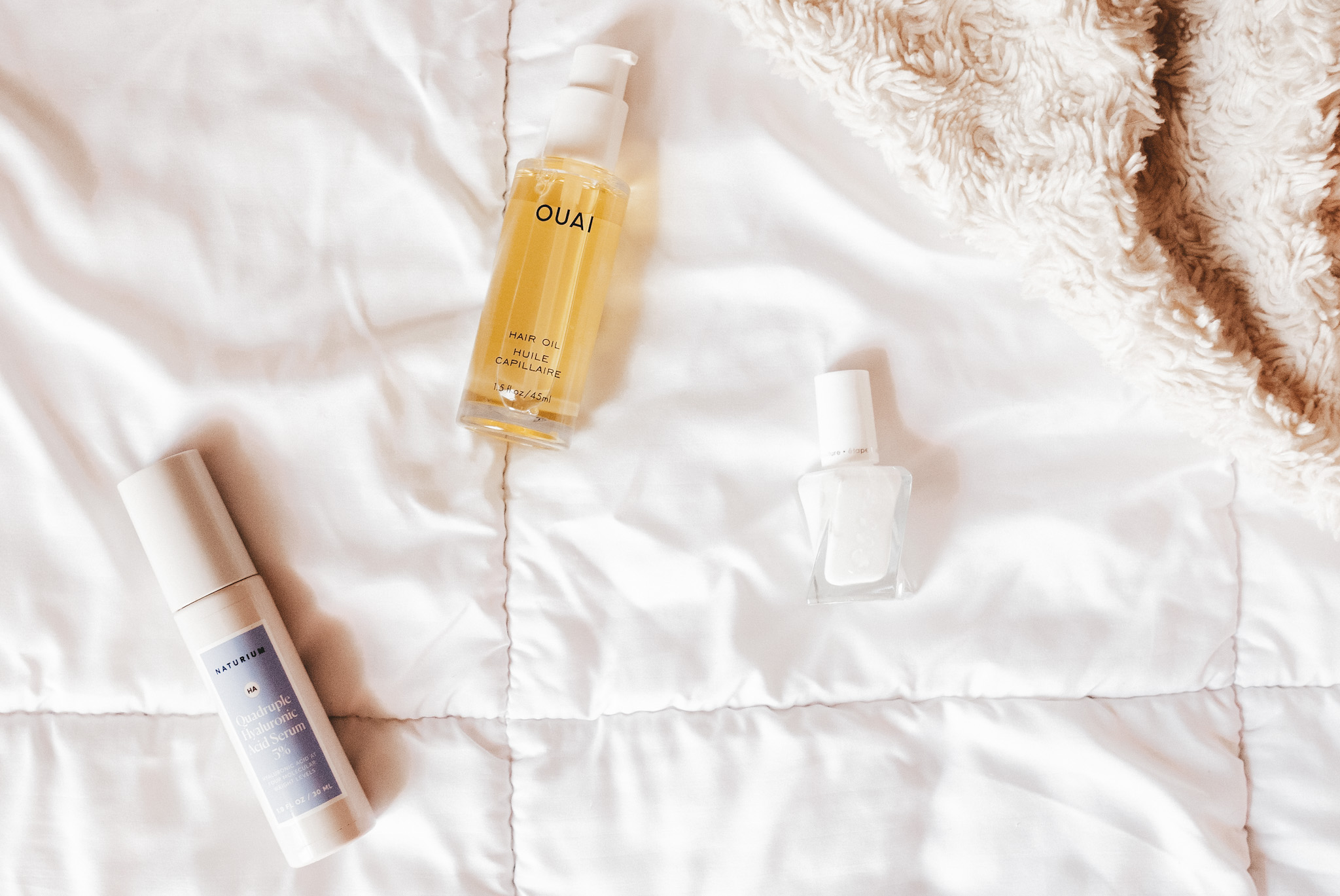 a white comforter, a bottle of Quai Hair Oil, Naturium Hyaluronic Acid and Essie Gel Couture Nail Polish