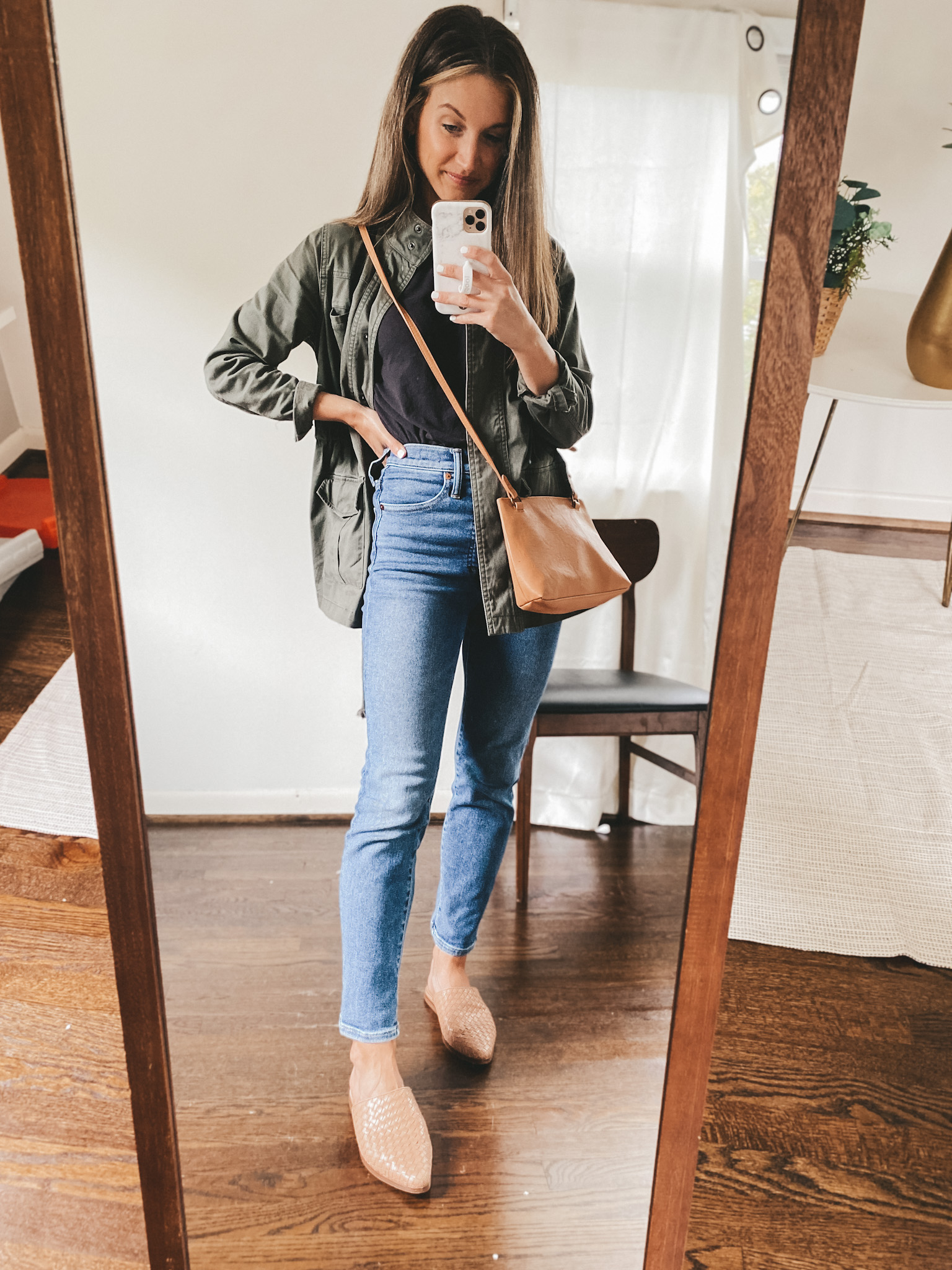 ashley wearing a black tee, green utility jacket, mid-wash denim and woven mules with a camel colored crossbody bag