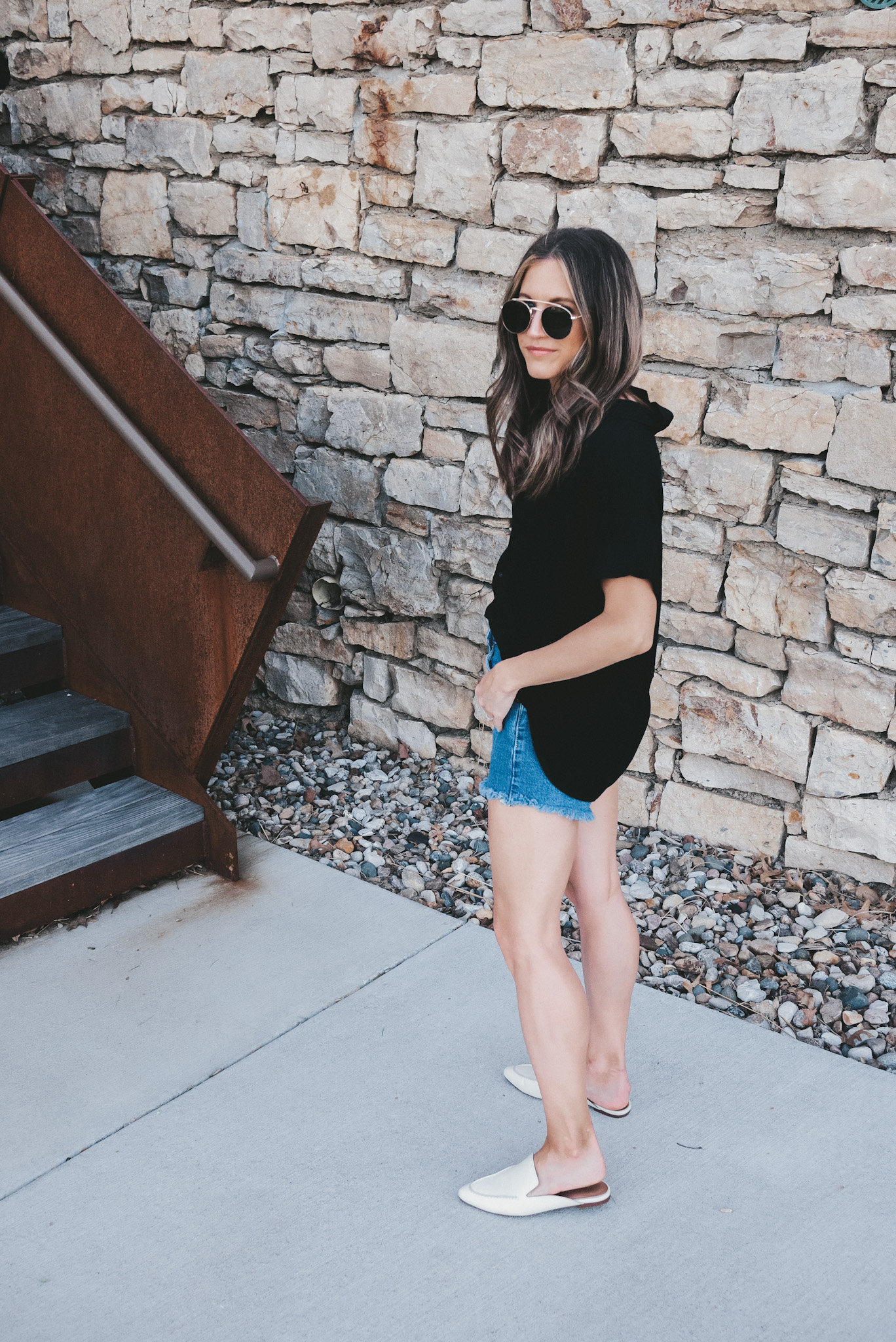 ashley wearing a black drapey button down top, cut-off denim shorts, white mules and circle frame sunglasses standing next to a stone wall