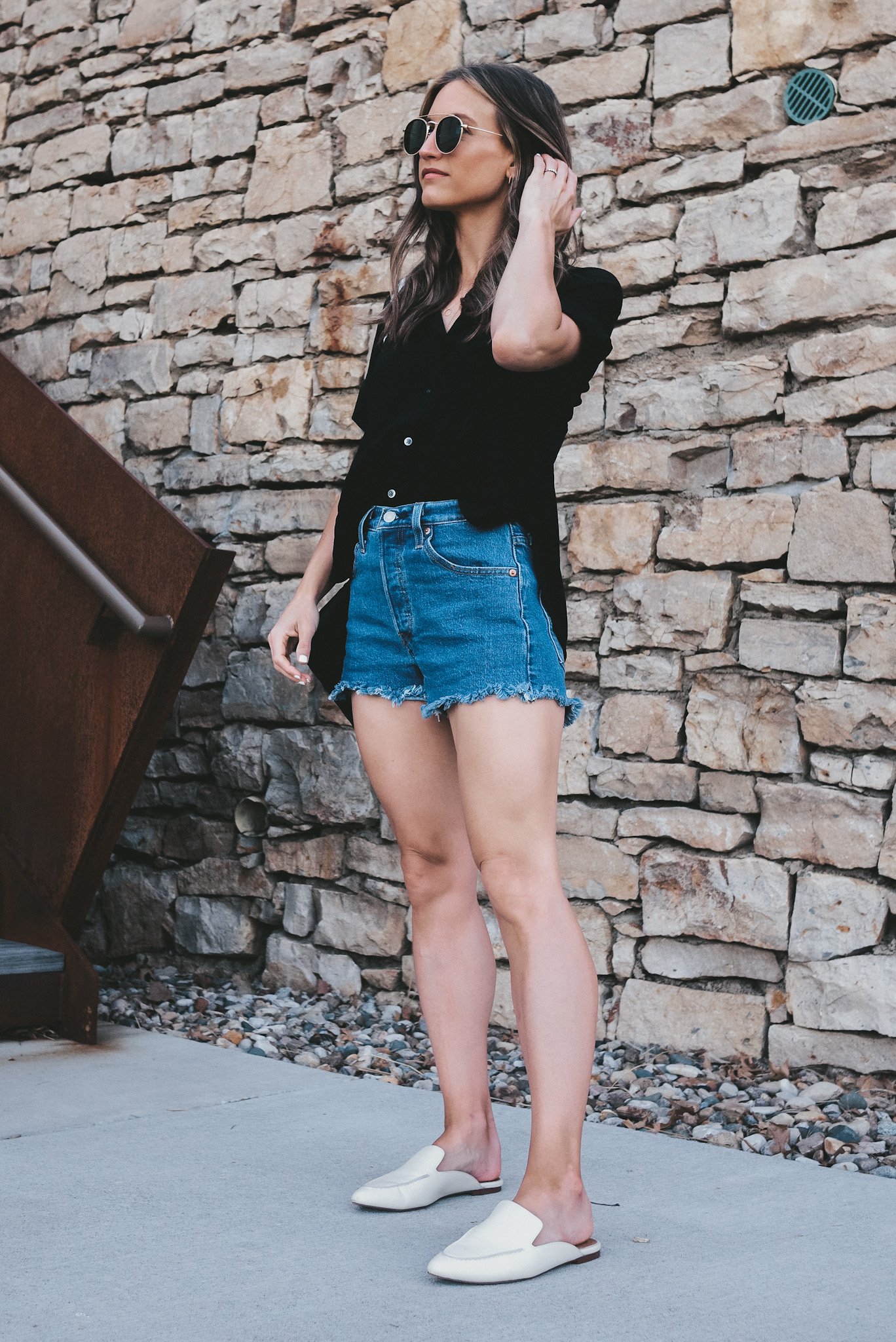 ashley wearing a black drapey button down shirt, mid-wash cut-off denim shorts, white mules and circle frame sunglasses standing next to a stone wall