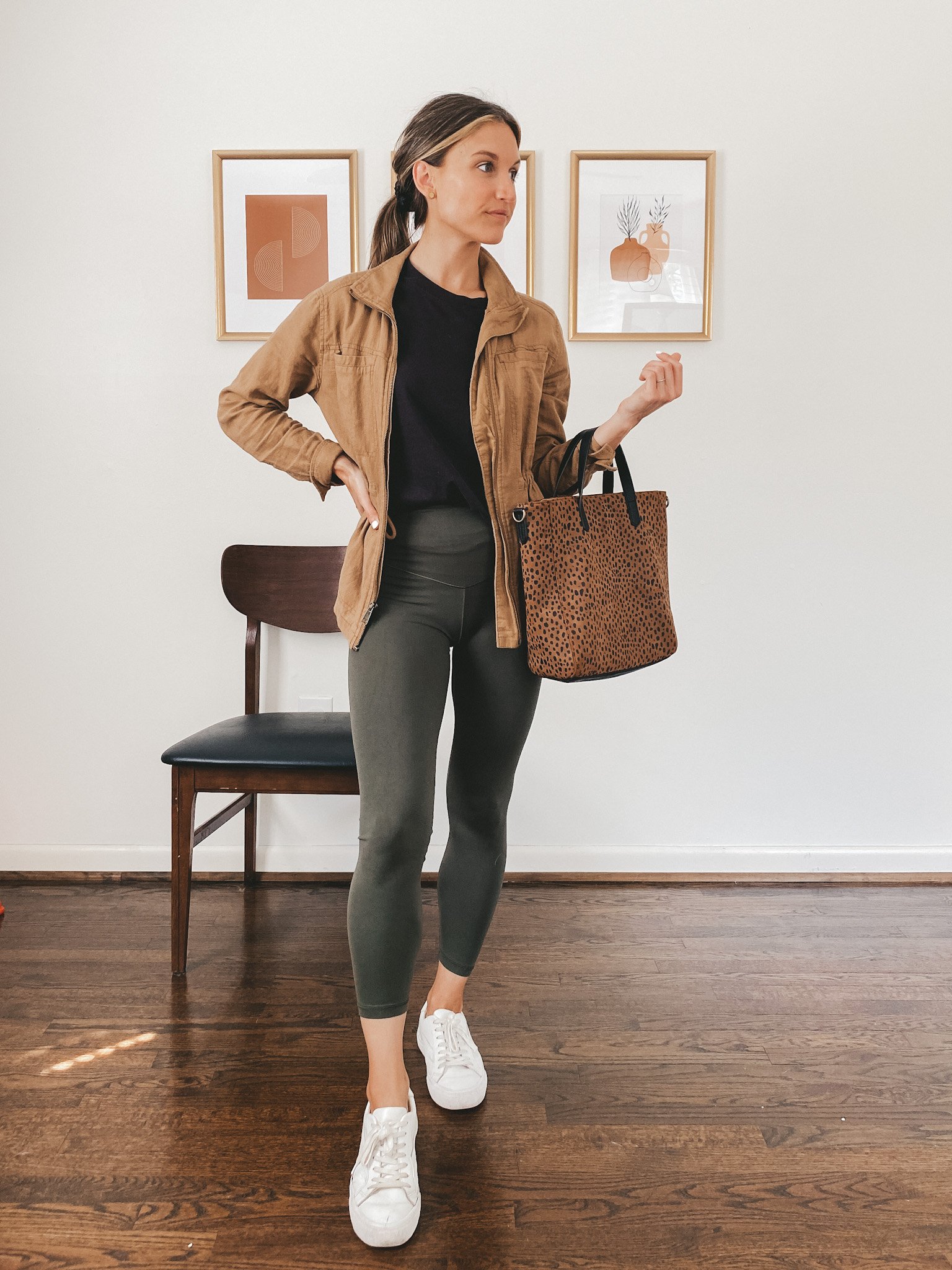 ashley wearing a black tee, tan utility jacket, olive green leggings and white sneakers carrying a leopard tote bag