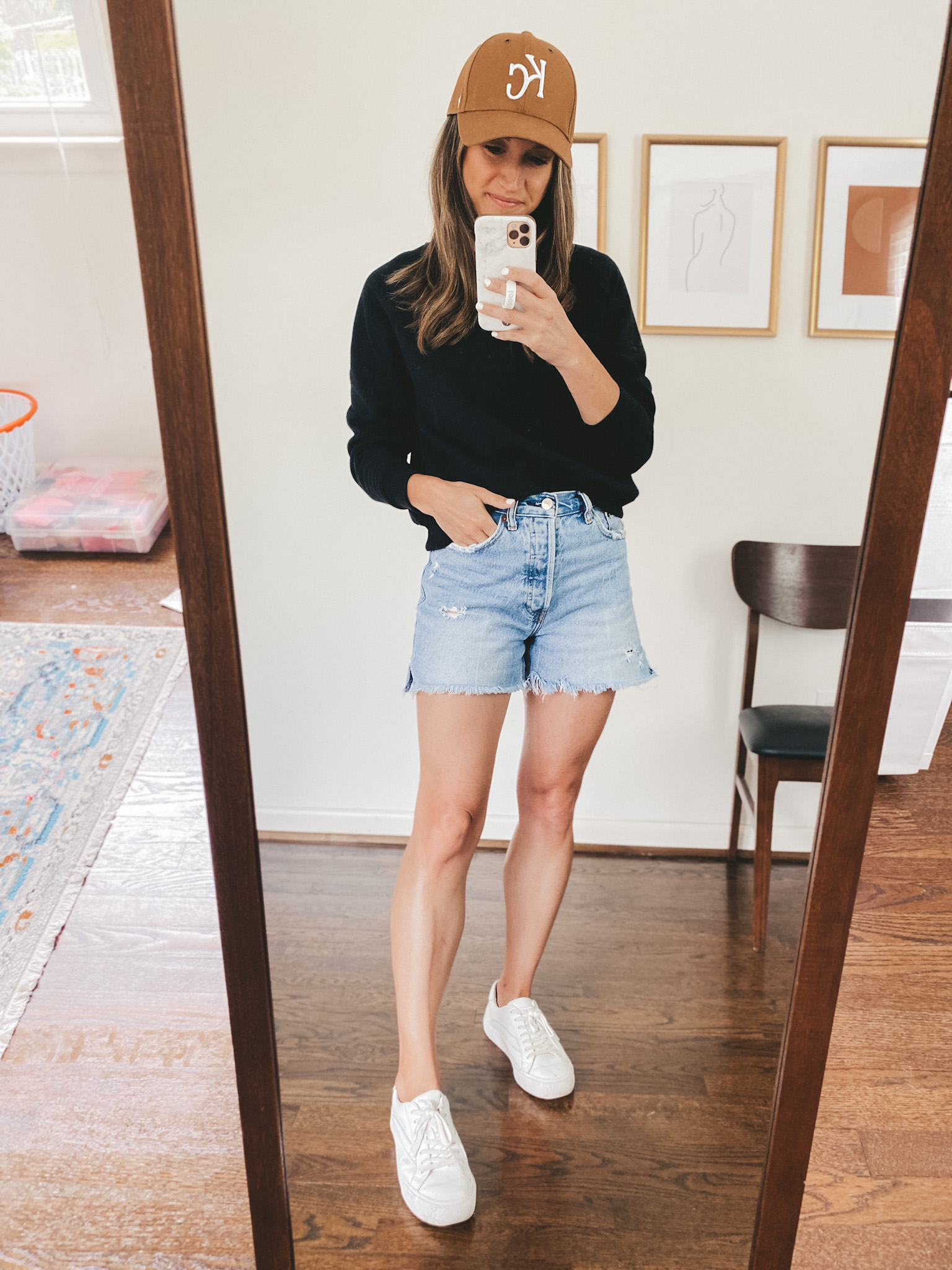 ashley wearing a black crewneck sweatshirt, denim cut-off shorts, white sneakers, and a brown KC baseball hat